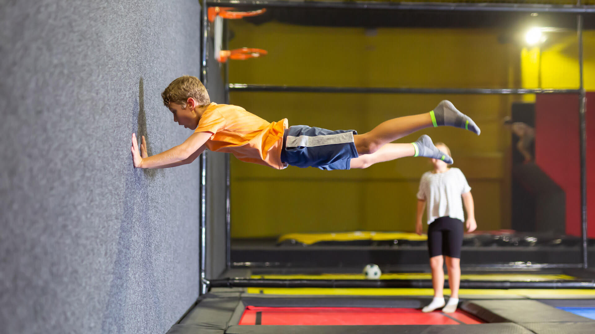Ein Junge in einem orangefarbenen T-Shirt und blauen Shorts springt in einem Trampolinpark und drückt sich mit beiden Händen gegen eine gepolsterte Wand. Er befindet sich in der Luft, waagerecht mit ausgestrecktem Körper, während seine Füße in grauen Socken vom Boden abgehoben sind. Im Hintergrund steht ein weiteres Kind in weiß gestreiftem Oberteil und schwarzen Shorts auf einem der Trampoline. Der Raum ist hell erleuchtet, und die Trampoline sind von Sicherheitsnetzen und gepolsterten Wänden umgeben. Foto: ideas4you.at