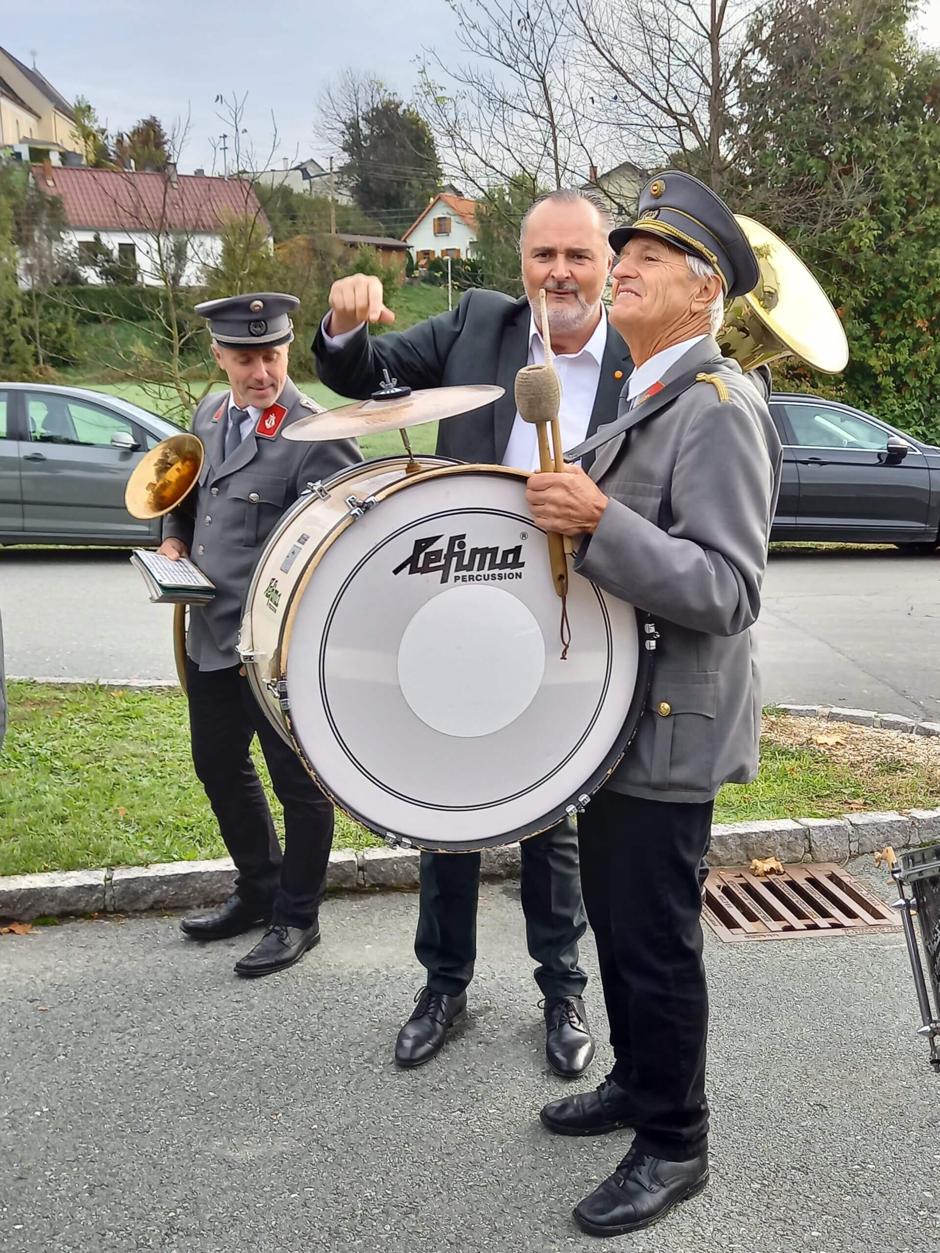 Ein Mann in schwarzem Anzug steht hinter einem uniformierten Mitglied einer Blaskapelle und schlägt spielerisch mit einem Schlegel auf eine große Bassdrum. Das Kapellenmitglied, das eine Schirmmütze trägt, lächelt und hält die Trommel fest. Ein weiteres Mitglied der Kapelle in grauer Uniform steht daneben und hält ein Blechblasinstrument sowie einen Notenhalter. Im Hintergrund sind Bäume, parkende Autos und Häuser mit roten Dächern zu sehen. Die Szene findet draußen auf einer asphaltierten Straße statt, die von Gras und einem niedrigen Bordstein gesäumt ist.