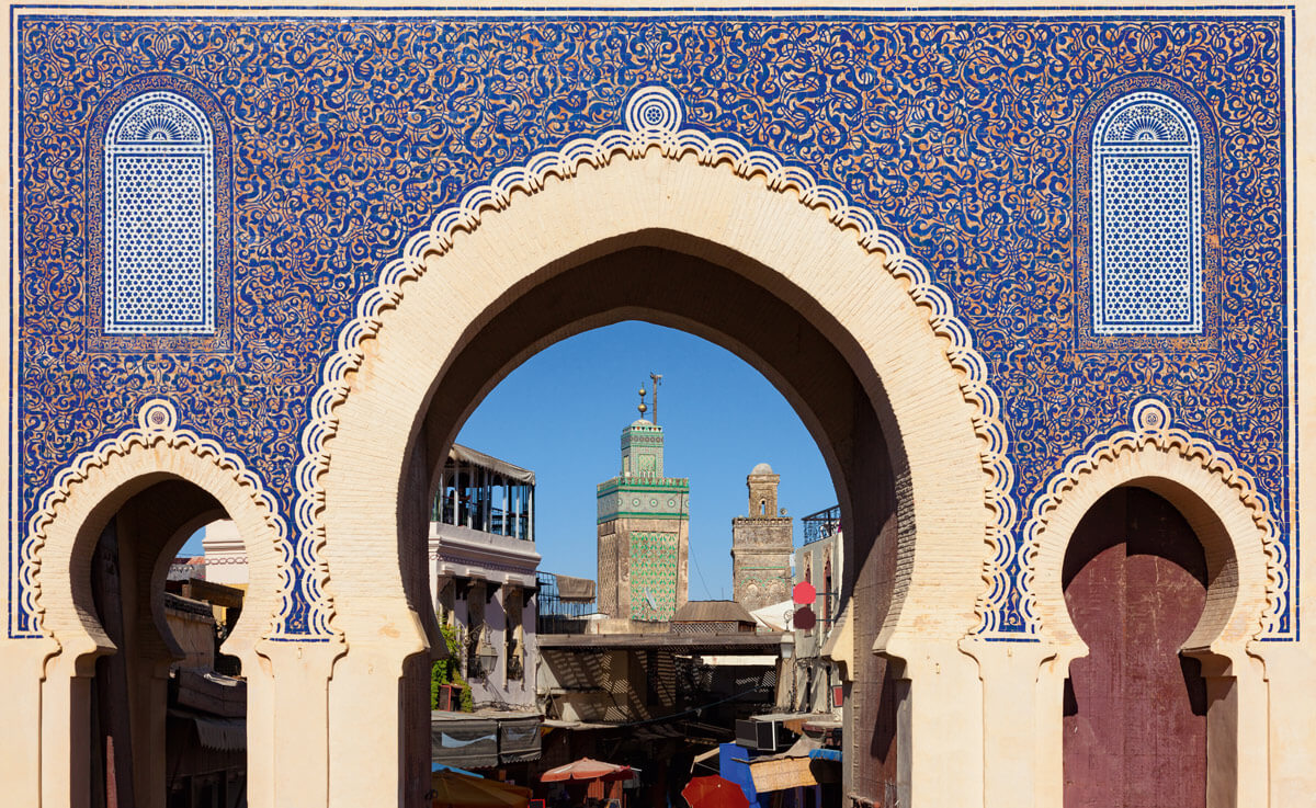 Ein kunstvoll verzierter Torbogen in Fès, bekannt als Bab Bou Jeloud, mit blauen Kacheln und weißen Mustern. Durch den Bogen ist ein Blick auf die Minarette und die Dächer der Altstadt zu sehen.