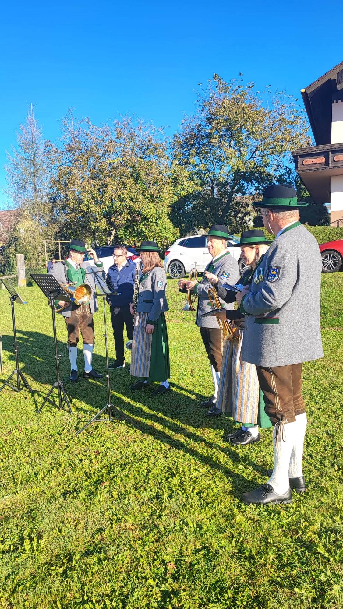 Eine kleine Gruppe von Musikanten in traditioneller Tracht steht auf einer Wiese, die von der Sonne beschienen wird. Sie tragen graue Janker, grüne Hüte mit Feder und spielen Blechblasinstrumente. Einige der Musiker sprechen miteinander, während sie ihre Instrumente halten. Im Hintergrund sind Bäume und parkende Autos zu sehen, und der Himmel ist strahlend blau