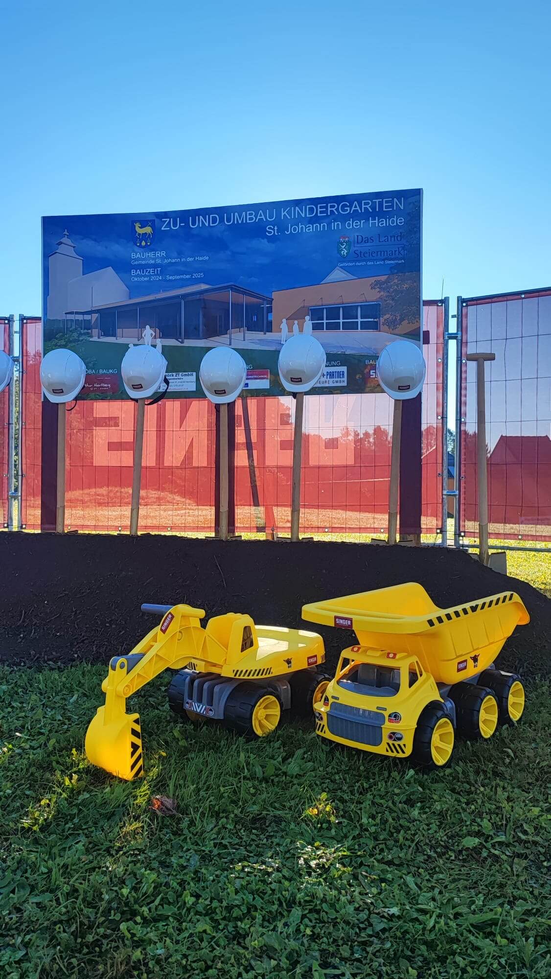 Das Foto zeigt zwei große gelbe Spielzeugfahrzeuge—einen Spielzeugbagger und einen Spielzeugkipper—auf einer Wiese vor einer Baustelle. Im Hintergrund ist ein Schild zu sehen mit der Aufschrift "Zu- und Umbau Kindergarten St. Johann in der Haide", das den geplanten Entwurf des neuen Kindergartengebäudes zeigt. Vor dem Schild sind mehrere weiße Bauhelme an Holzpfosten aufgehängt. Der Himmel ist klar und sonnig.
