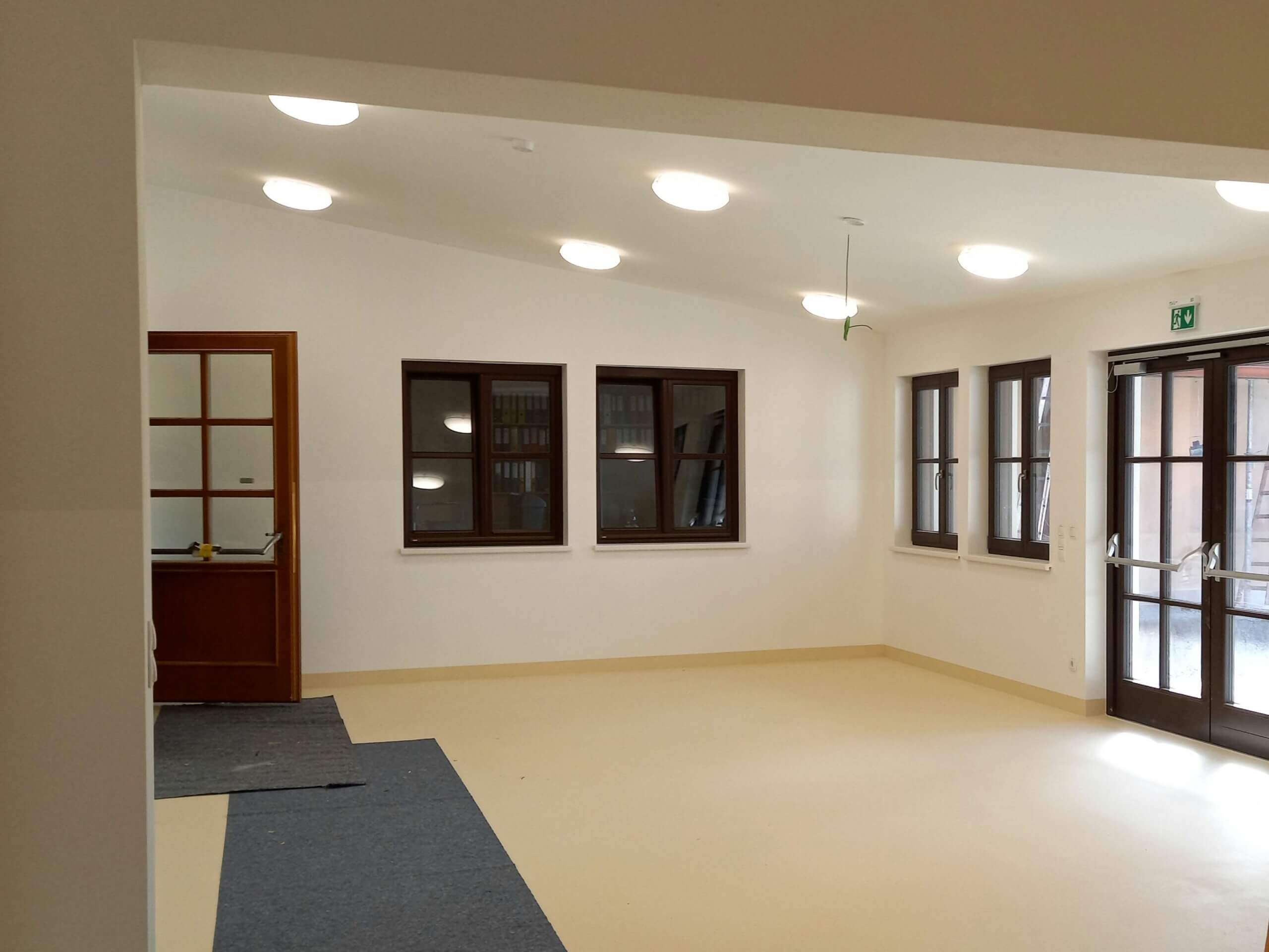 Ein leerer, lichtdurchfluteter Raum mit cremefarbenem Boden und weißen Wänden. Links führt eine Holztür zu einem angrenzenden Bereich, und rechts befindet sich eine große Fensterfront mit Glastüren, die nach draußen führt. An der Decke sind mehrere runde Deckenleuchten angebracht.