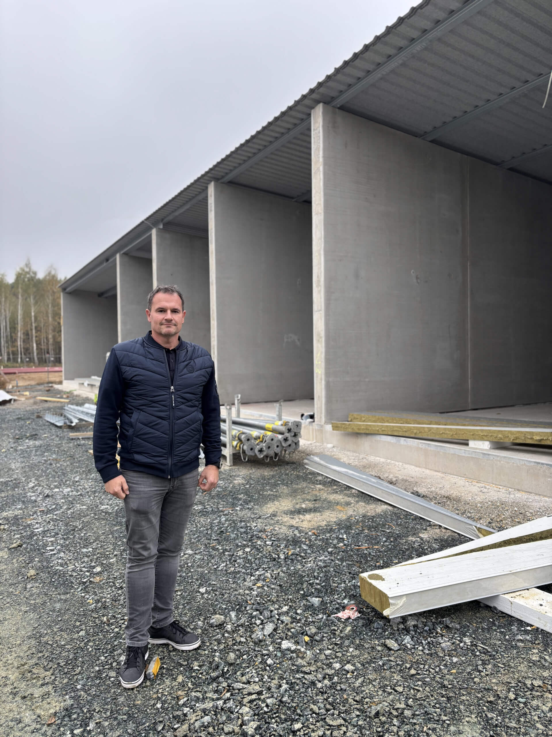 Bürgermeister Wolfgang Koller steht vor dem Rohbau mit überdachten Betonabschnitten am Bauhof Kemeten. Der Boden ist mit Schotter bedeckt, und im Hintergrund sind Bäume zu sehen. Der Mann trägt eine dunkle Jacke und Jeans.