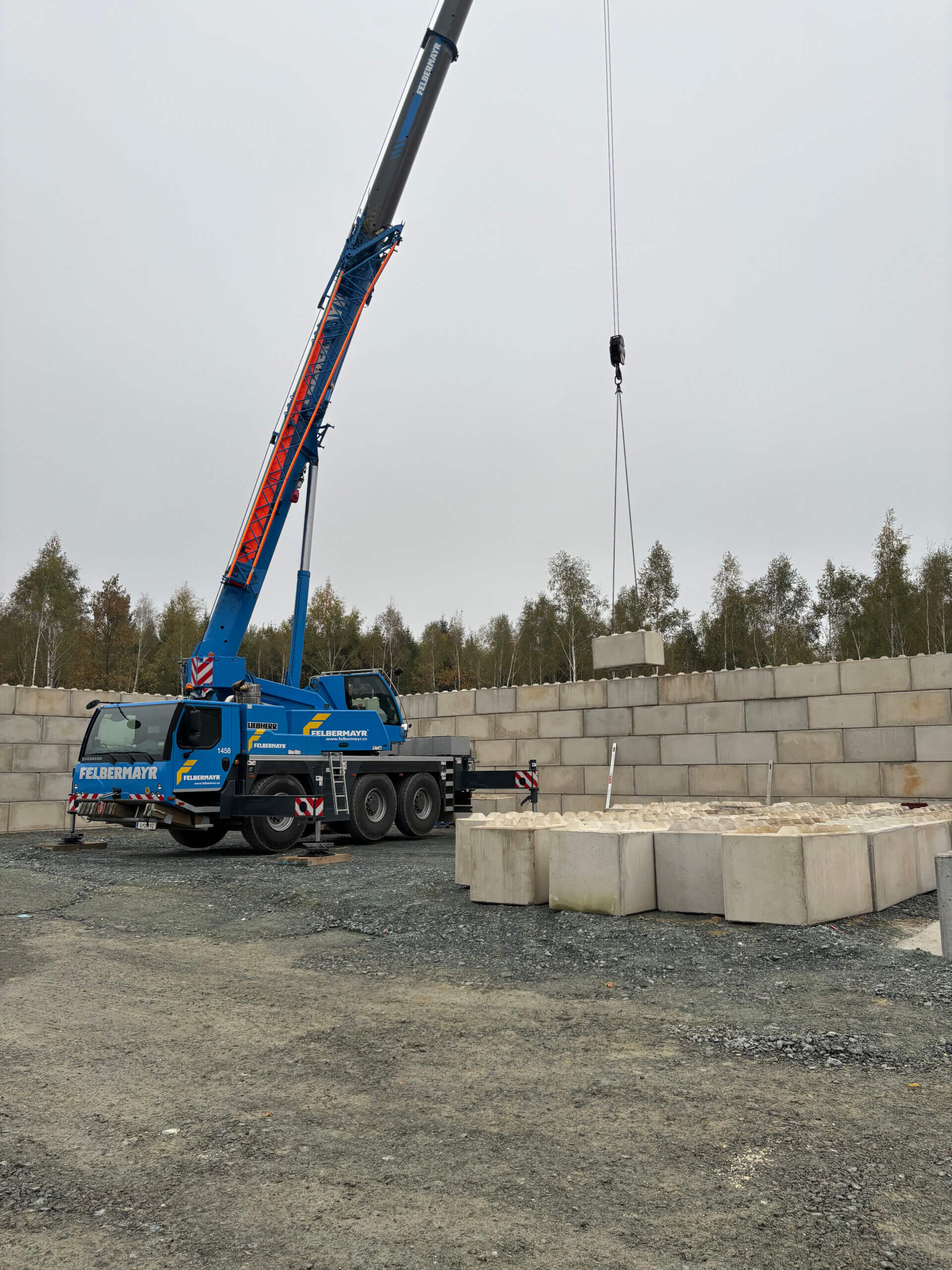 Ein großer blauer Kran von "Felbermayr" hebt Betonblöcke auf dem Bauhof Kemeten. Die Baustelle ist von Stapeln großer Betonsteine umgeben, und im Hintergrund sind Bäume und ein grauer Himmel zu sehen.