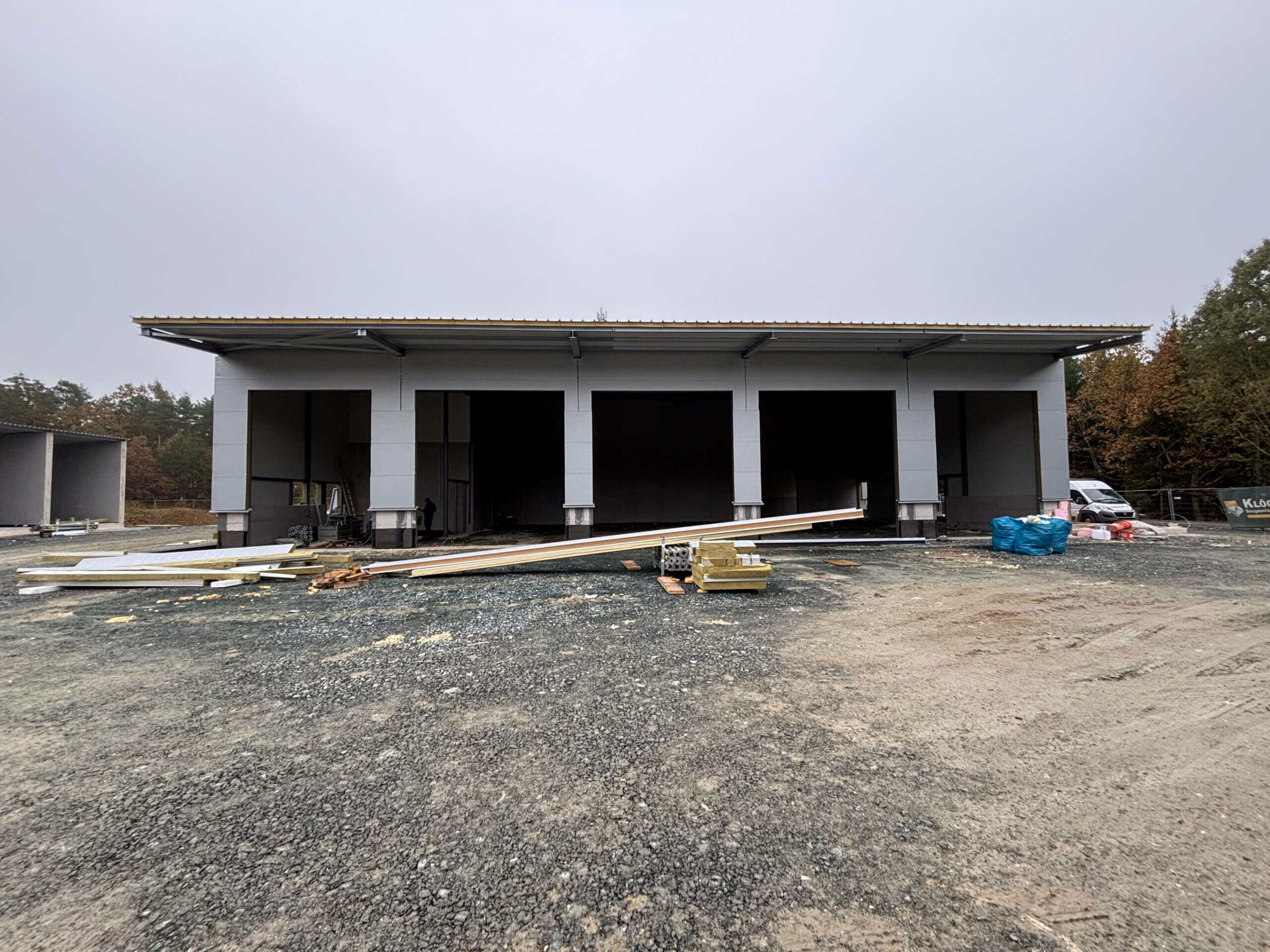 Frontansicht des Bauhof-Gebäudes im Bauzustand. Das Gebäude hat offene Eingänge ohne Türen, und Baumaterialien liegen im Vordergrund auf dem Schotterboden verteilt.