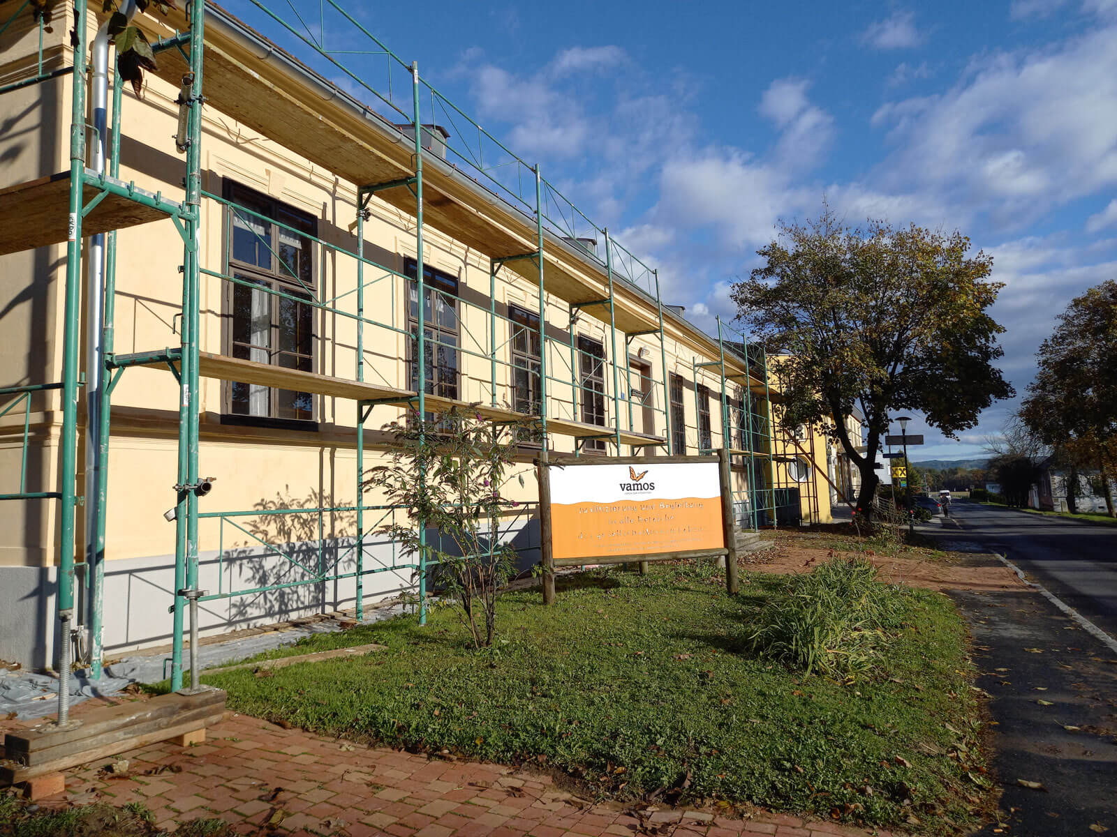 Außenansicht eines Gebäudes mit Gerüst an der Fassade. Die cremefarbene Wand hat große, dunkle Fenster. Vor dem Gebäude steht ein Schild mit dem Logo "Vamos". Rechts im Bild ist eine Straße zu sehen, im Hintergrund ein Baum mit Herbstlaub.