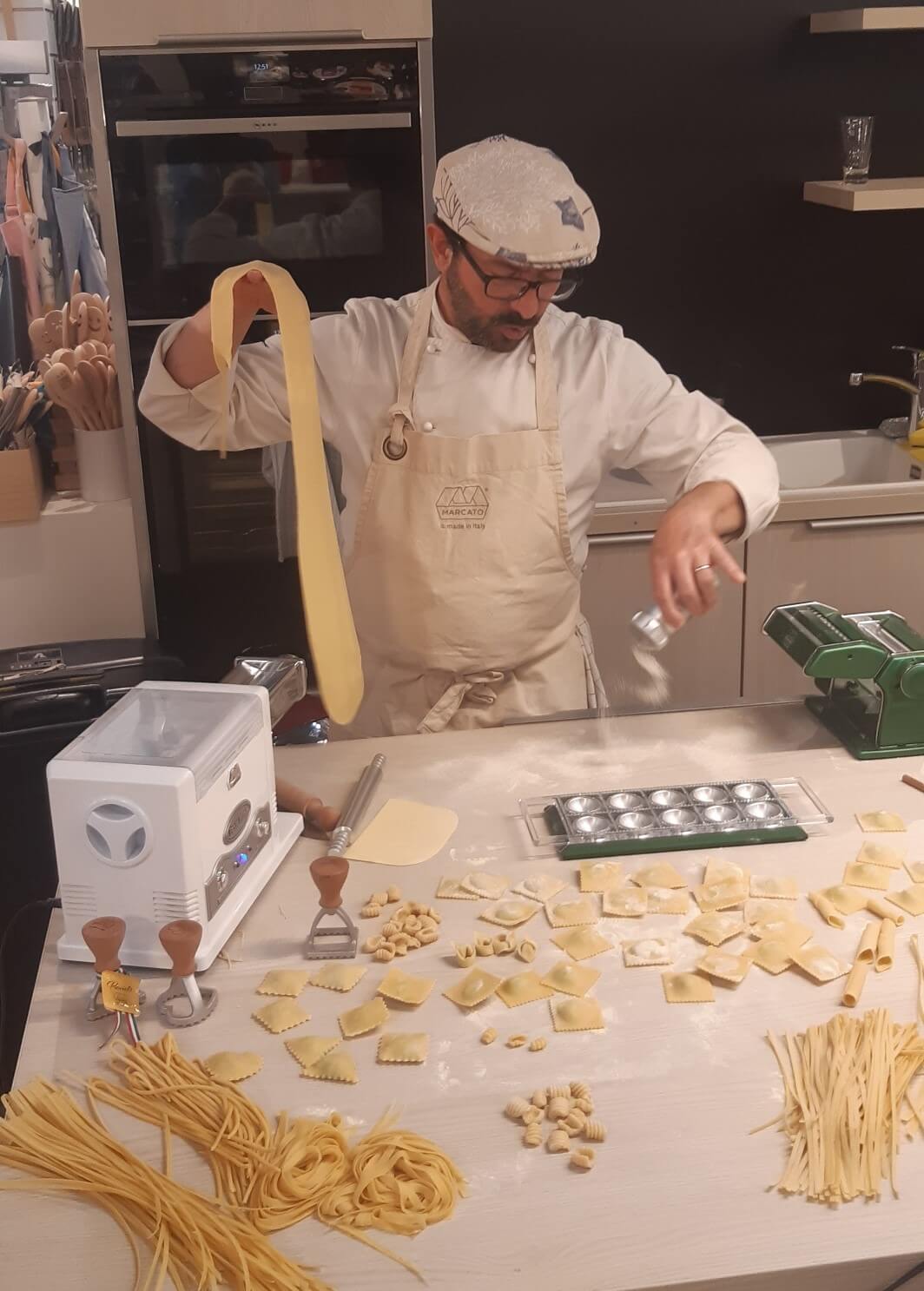 Ein italienischer Koch mit Brille und Mütze arbeitet an einem langen Teigstreifen. Vor ihm liegt eine Vielzahl frisch hergestellter Pastasorten, darunter Ravioli, Spaghetti und Fusilli, die ordentlich auf der Arbeitsfläche ausgebreitet sind. Er trägt eine Schürze und streut vorsichtig Mehl auf die Pasta.