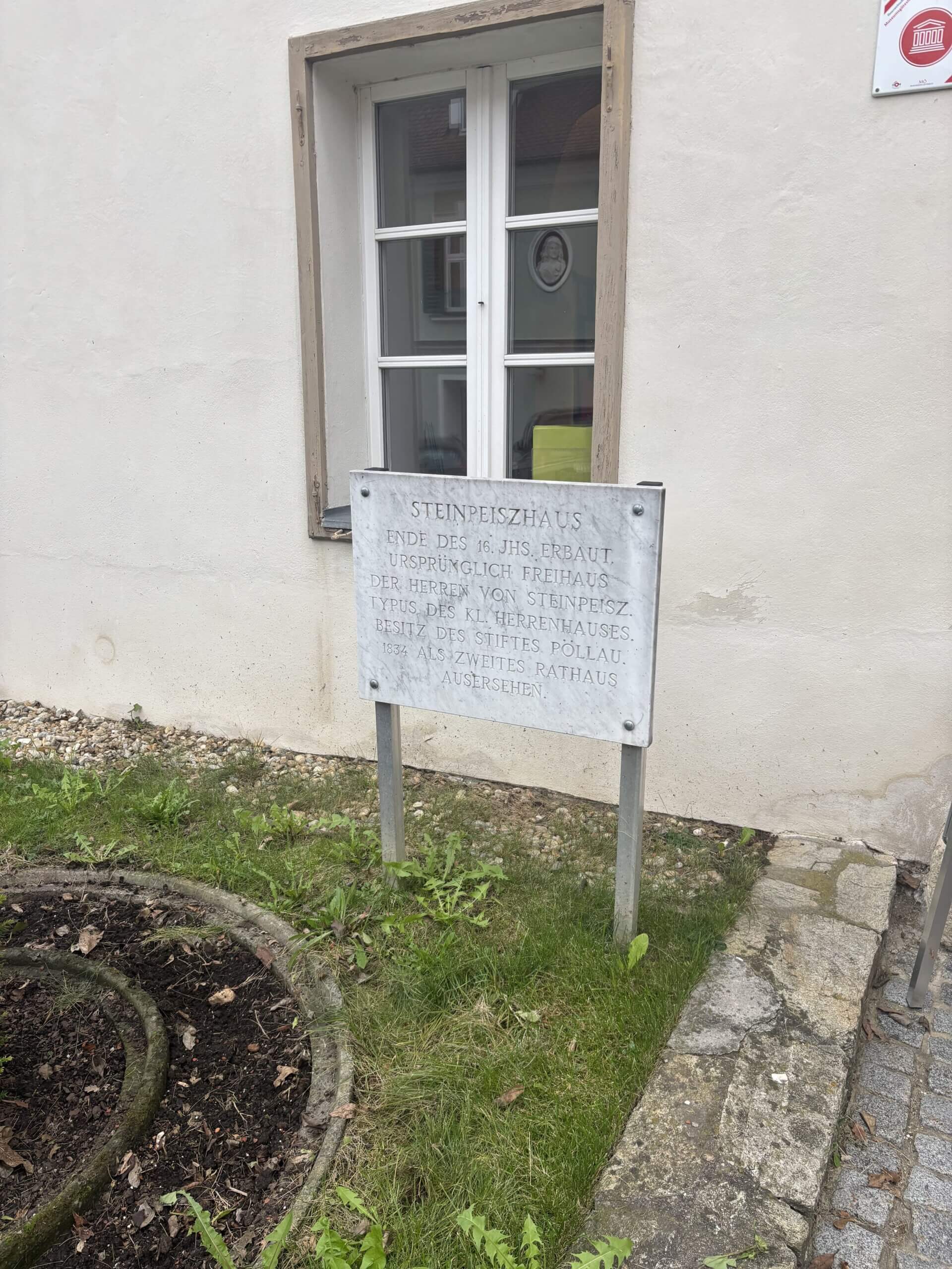 Nahaufnahme einer Informationstafel neben einem Fenster des Steinpeißhauses. Die Tafel beschreibt das Gebäude und seine historische Bedeutung und steht neben einer kleinen, kreisförmigen Beetfläche.