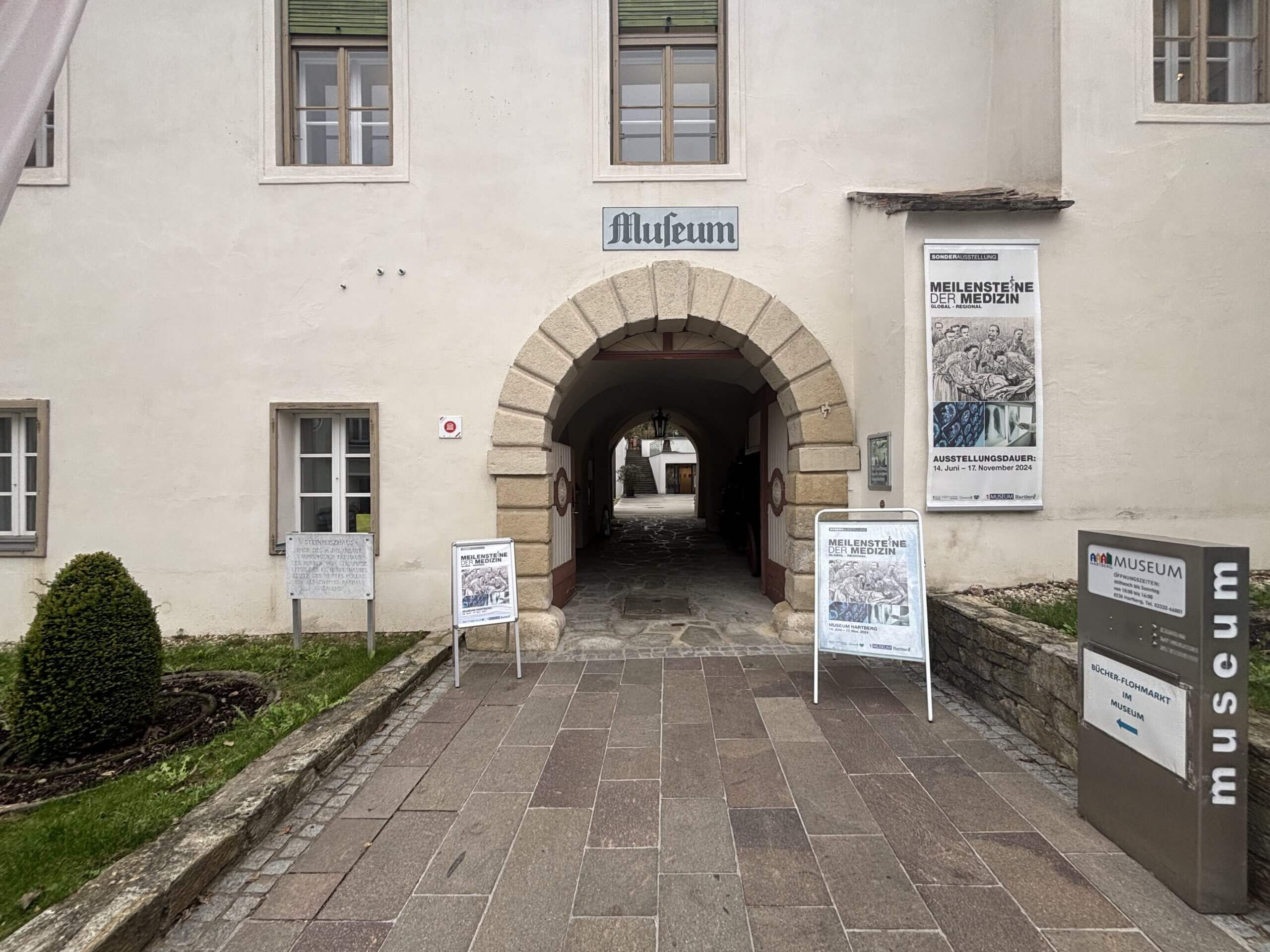 Der Eingang des Museums im Steinpeißhaus, ein großer Torbogen aus Stein mit der Aufschrift "Museum". Links und rechts des Eingangs stehen Informationstafeln zur aktuellen Ausstellung, und eine kleine Grünfläche mit einem Beet ist zu sehen.