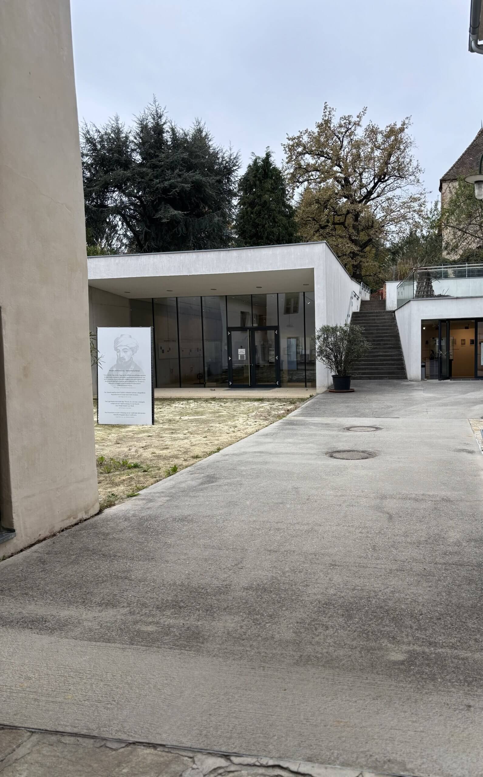 Ein Blick in den modernen Anbau des Steinpeißhauses mit großen Glasfenstern und einer weiteren Tafel mit einer Illustration eines Mannes im Innenhof. Eine Treppe führt rechts am Gebäude entlang.