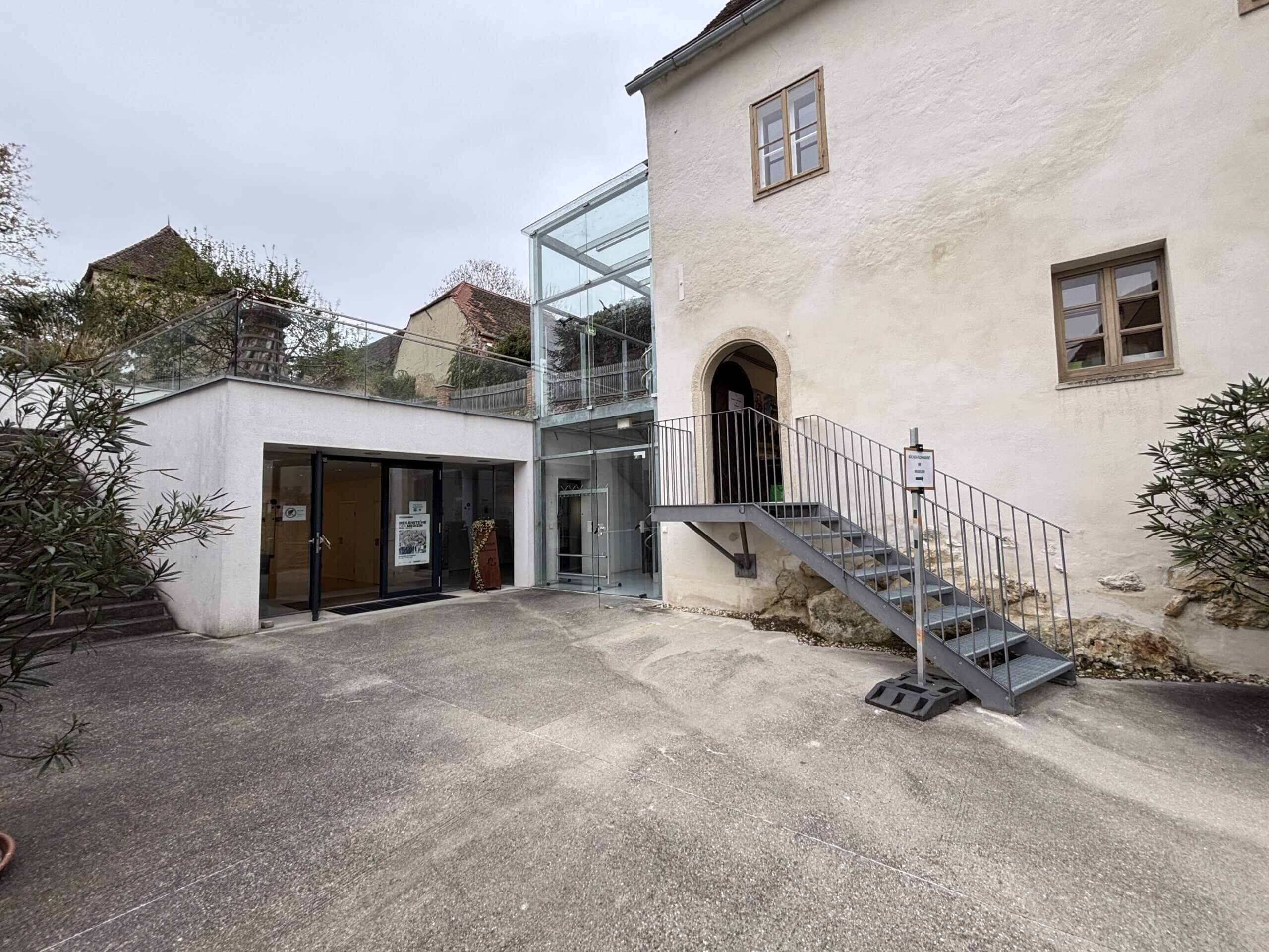 Eine Außenaufnahme des modernen Teils des Steinpeißhauses mit einer großen Glasfront und einer Treppe, die auf eine Terrasse führt. Rechts ist eine kleine Pflanze im Topf.