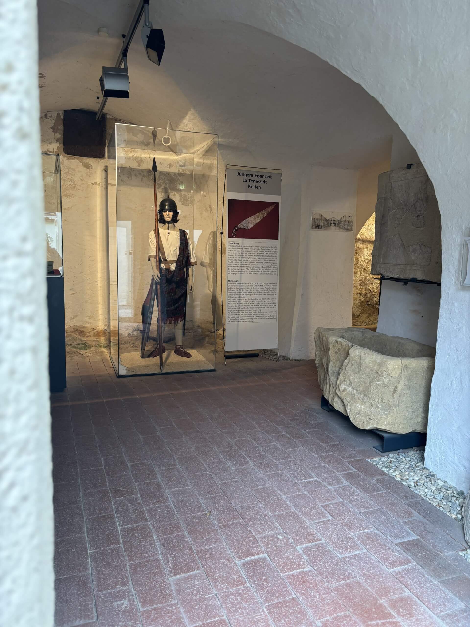 Innenaufnahme eines Ausstellungsraums im Steinpeißhaus. In einem Glasgehäuse steht eine lebensgroße Figur eines Kriegers in historischer Kleidung, daneben Informationstafeln zu Ausstellungsobjekten.