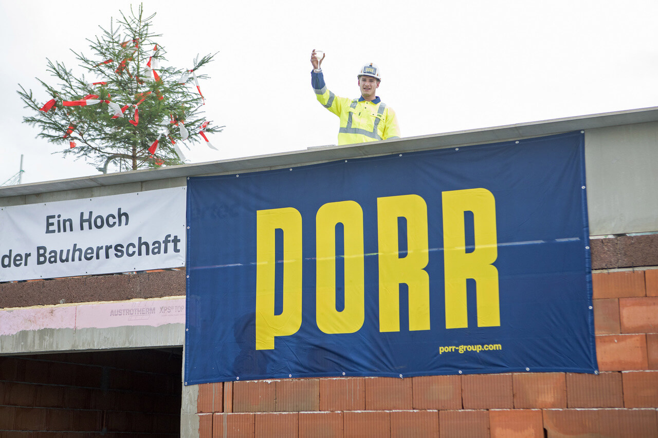 Ein Bauarbeiter steht auf dem Dach eines Rohbaus und hebt ein Glas zur Feier der Dachgleiche. Er trägt eine Schutzweste und einen Helm. Im Hintergrund ist ein Richtbaum mit roten und weißen Bändern zu sehen. Unter ihm hängt ein großes Banner des Bauunternehmens "PORR". Daneben ist ein weiteres Banner mit der Aufschrift „Ein Hoch der Bauherrschaft“ angebracht.