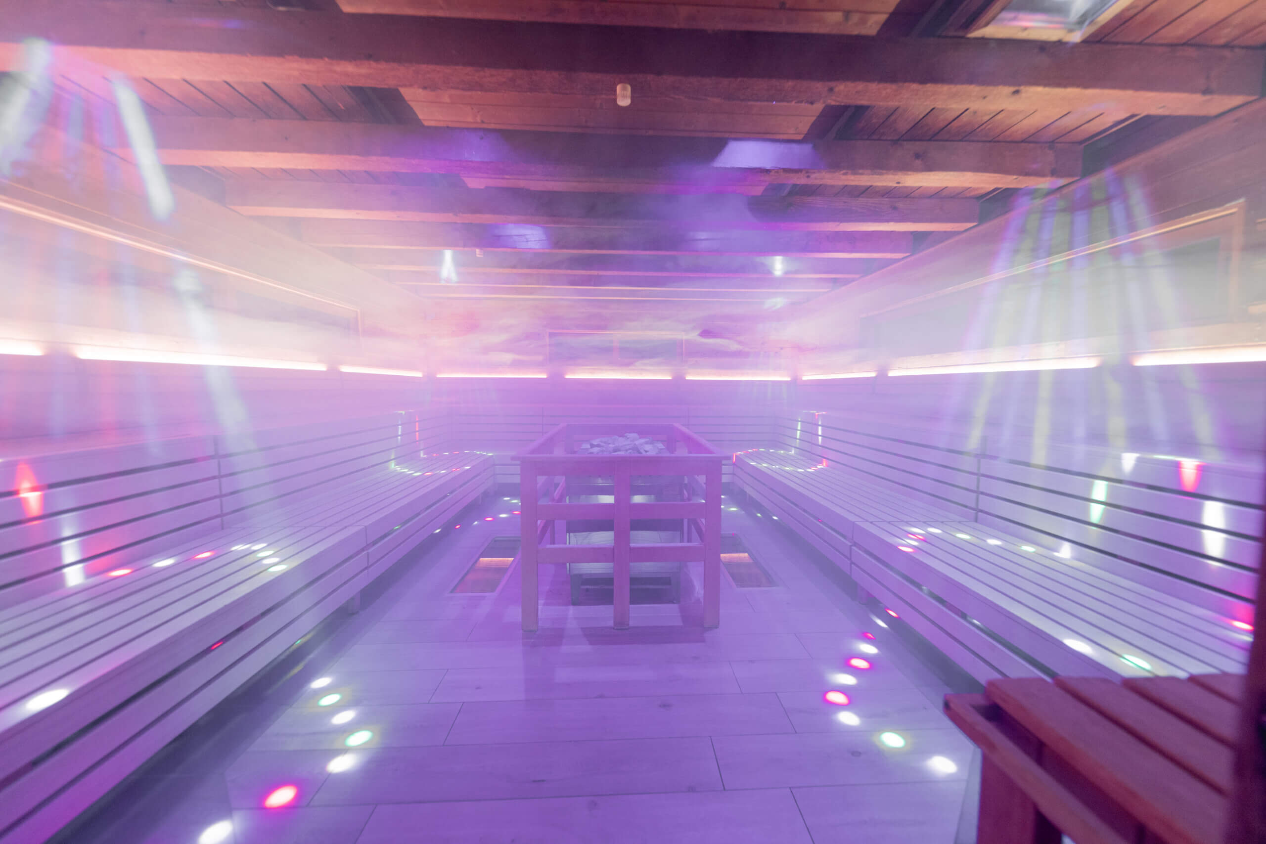 Eine modern gestaltete Sauna im AVITA Resort mit einer stimmungsvollen Lichter- und Nebelinstallation. Die Sitzbänke verlaufen entlang der Wände, und im Zentrum befindet sich ein Saunaofen. Das Lichtspiel mit farbigen Akzenten und der Dampf verleihen dem Raum eine besondere Atmosphäre, die Entspannung und Erlebnis vereint.