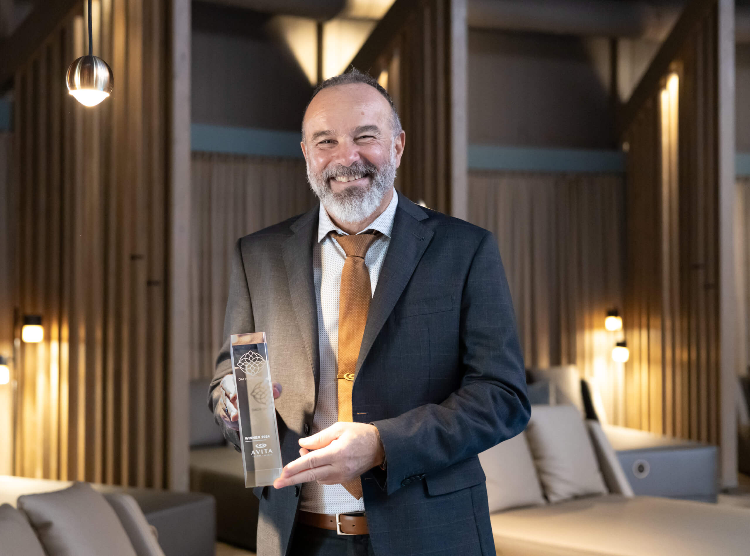 Peter Prisching, Geschäftsführer des AVITA Resorts, hält lächelnd die Trophäe des DACH Spa Awards. Er trägt einen Anzug und steht in einer modernen, eleganten Umgebung des Resorts, die durch warmes Licht und stilvolle Dekoration geprägt ist. Im Hintergrund sind weitere Lounge-Möbel sichtbar.