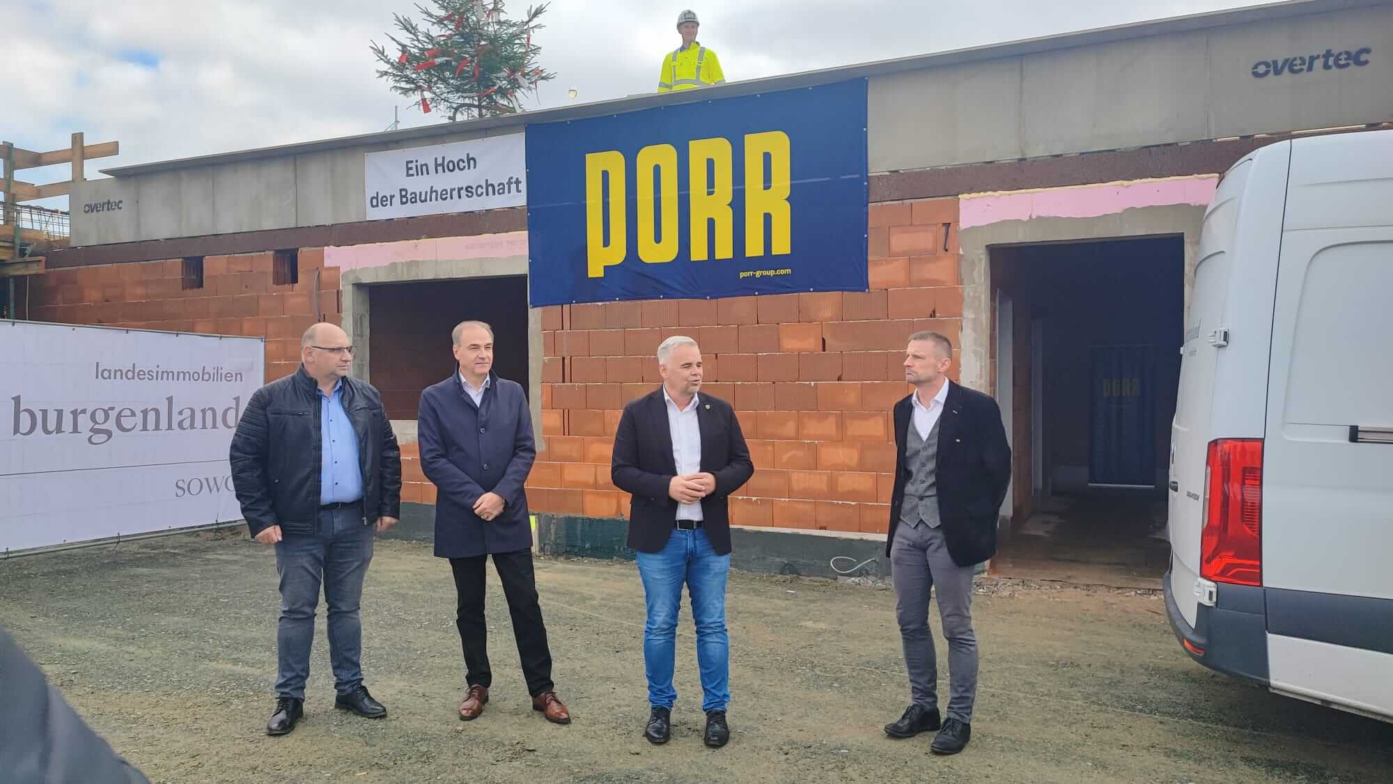 Vier Männer stehen vor einem Rohbau, der mit einem Banner des Bauunternehmens "PORR" versehen ist. Einer der Männer spricht, während die anderen zuhören. Über dem Eingang des Gebäudes ist ein Banner mit der Aufschrift „Ein Hoch der Bauherrschaft“ angebracht. Auf dem Dach steht ein Bauarbeiter mit Helm und Schutzweste, daneben ist der Richtbaum mit roten und weißen Bändern zu sehen.