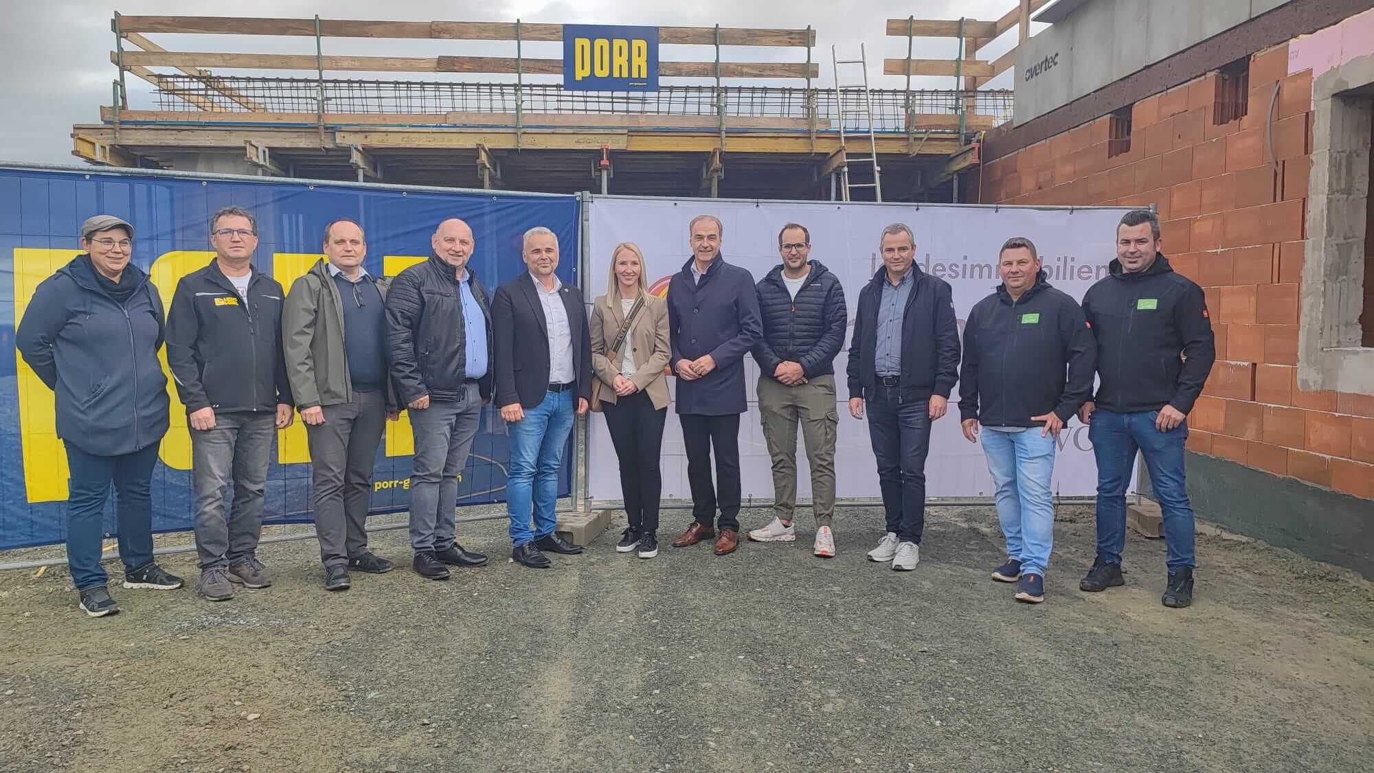 Elf Personen stehen vor einem Bauzaun, hinter dem eine Baustelle zu sehen ist. Alle blicken in die Kamera. Im Hintergrund ist ein teilweise fertiggestelltes Gebäude mit einem Banner des Bauunternehmens „PORR“ zu sehen. Die Gruppe trägt überwiegend Jacken und festes Schuhwerk, das Wetter wirkt kühl. Der Baufortschritt im Hintergrund zeigt Gerüste und Ziegelwände.