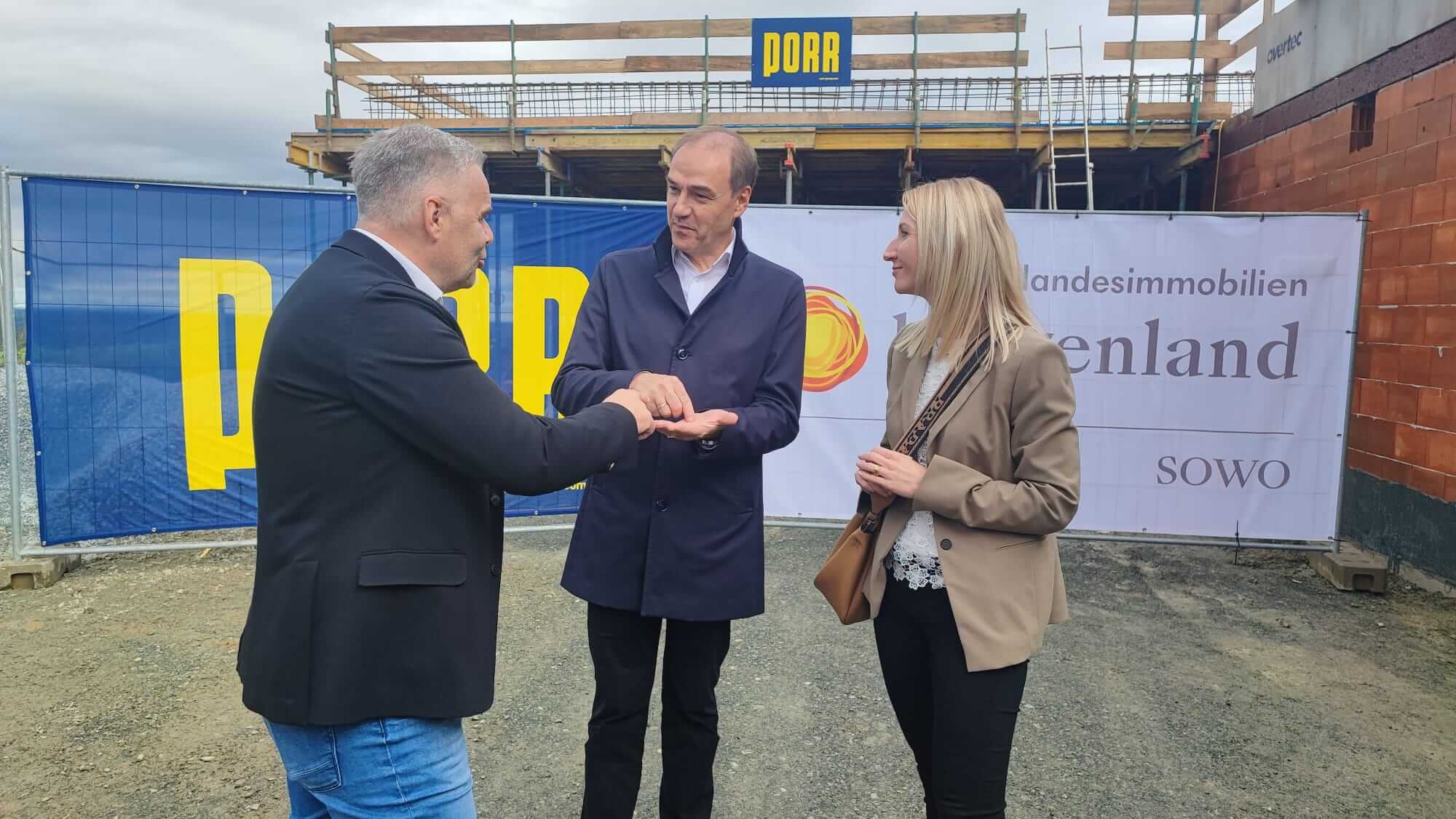 Drei Personen stehen vor einer Baustelle und unterhalten sich. Links ist ein Mann in schwarzem Sakko, in der Mitte ein Mann in einem dunklen Mantel, und rechts eine Frau im beigen Blazer. Im Hintergrund ist ein Bauzaun mit den Logos von „PORR“ und „Landesimmobilien Burgenland“ zu sehen. Hinter ihnen ist der Rohbau des Pflegestützpunkts zu erkennen.