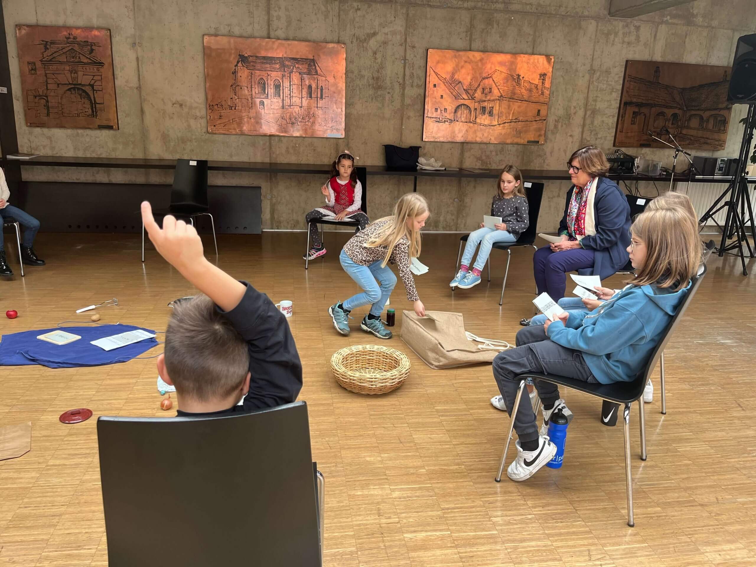 Eine Gruppe von sieben Kindern sitzt in einem Kreis auf schwarzen Stühlen in einem Raum mit Betonwänden und Holzboden. Ein Kind in der Mitte hebt einen Jutesack auf, während ein anderes Kind mit erhobenem Arm auf einem Stuhl im Vordergrund sitzt. Auf dem Boden liegen ein blaues Tuch, ein Korb und mehrere kleine Gegenstände. An der Wand hängen Kupfertafeln mit Abbildungen historischer Gebäude.