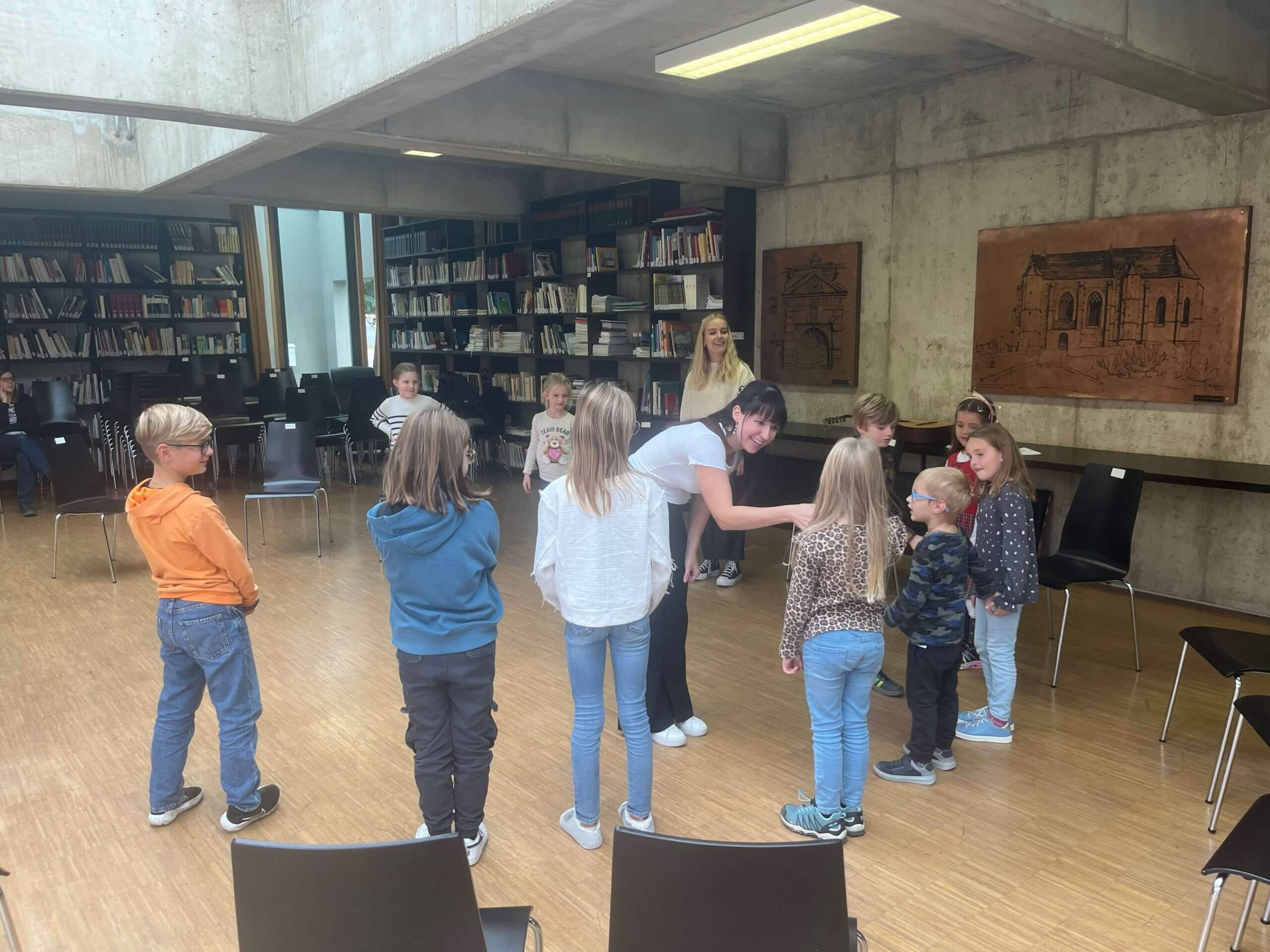 Mehrere Kinder stehen in einem Halbkreis in einem Raum mit hohen Betonwänden und großen Bücherregalen im Hintergrund. Zwei Erwachsene, eine von ihnen in weißem T-Shirt, interagieren mit den Kindern. An der Wand sind ebenfalls Kupfertafeln mit Gebäudemotiven befestigt. Die Stimmung wirkt entspannt und spielerisch.