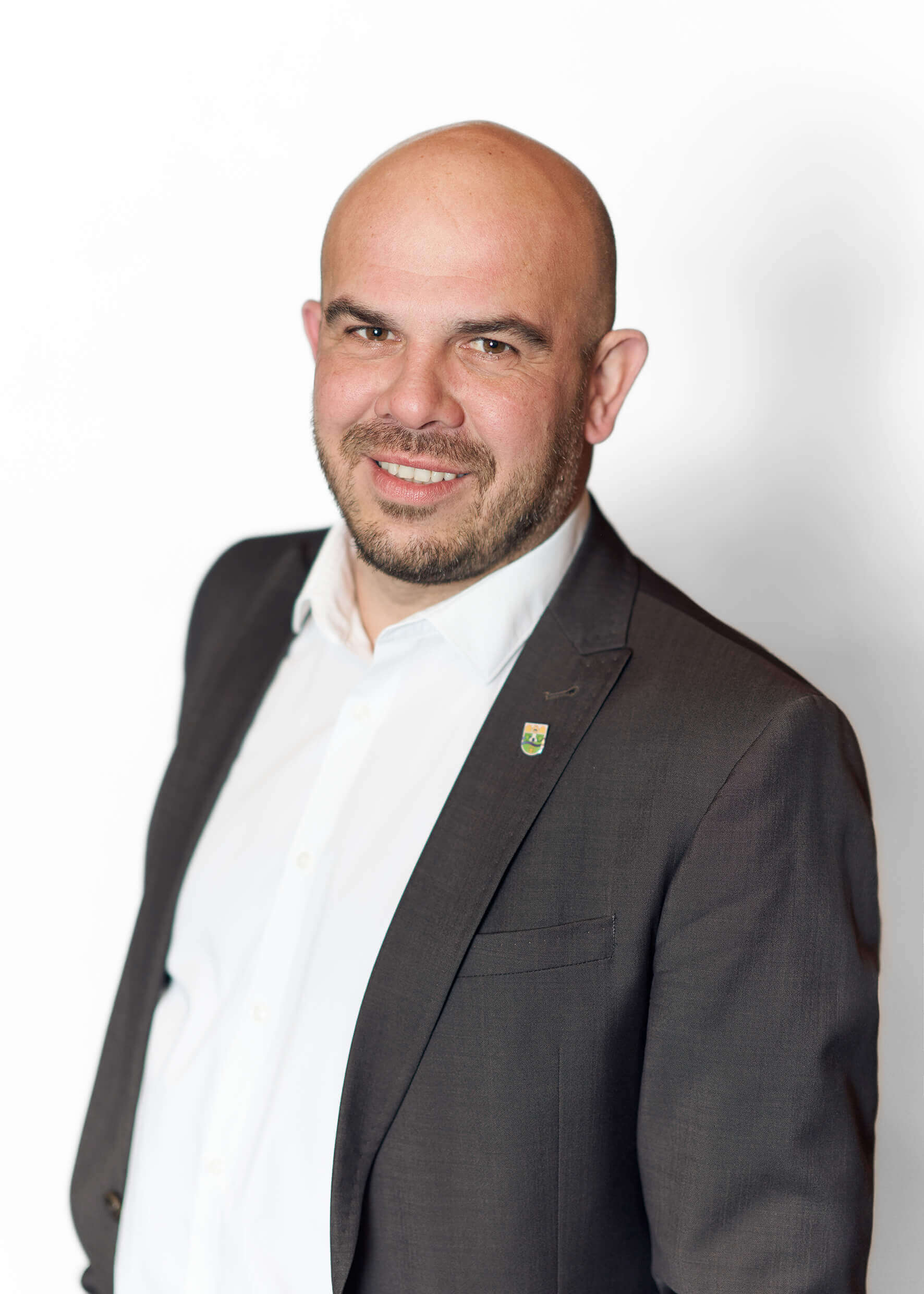 Joachim Raser, ein Mann mittleren Alters mit Glatze und Bart, in weißem Hemd und dunkelgrauem Sakko mit Anstecknadel, lächelnd vor weißem Hintergrund.