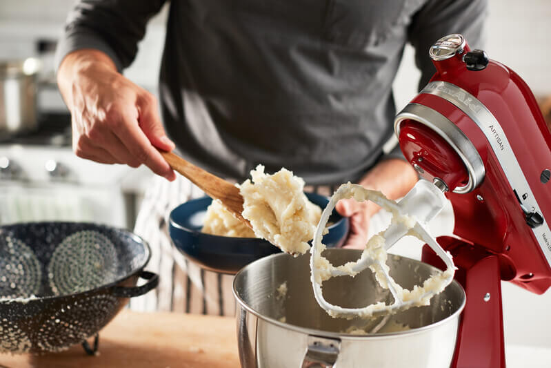 Eine Person schöpft mit einem Holzlöffel eine Portion Kartoffelpüree aus einer Schüssel. Daneben steht eine rote Küchenmaschine mit einem Rührbesen, die zur Zubereitung verwendet wurde.
