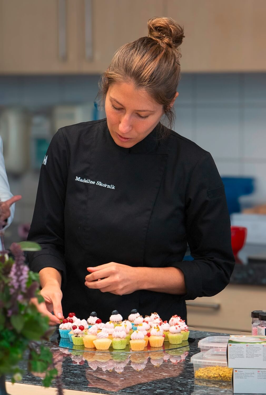 Konditorin Madeline steht an einem Tisch und dekoriert sorgfältig eine Auswahl an Mini-Cupcakes. Sie trägt eine schwarze Konditorjacke mit ihrem Namen darauf und konzentriert sich auf ihre Arbeit. Die Cupcakes sind in verschiedenen bunten Förmchen und mit Sahne und Beeren dekoriert.