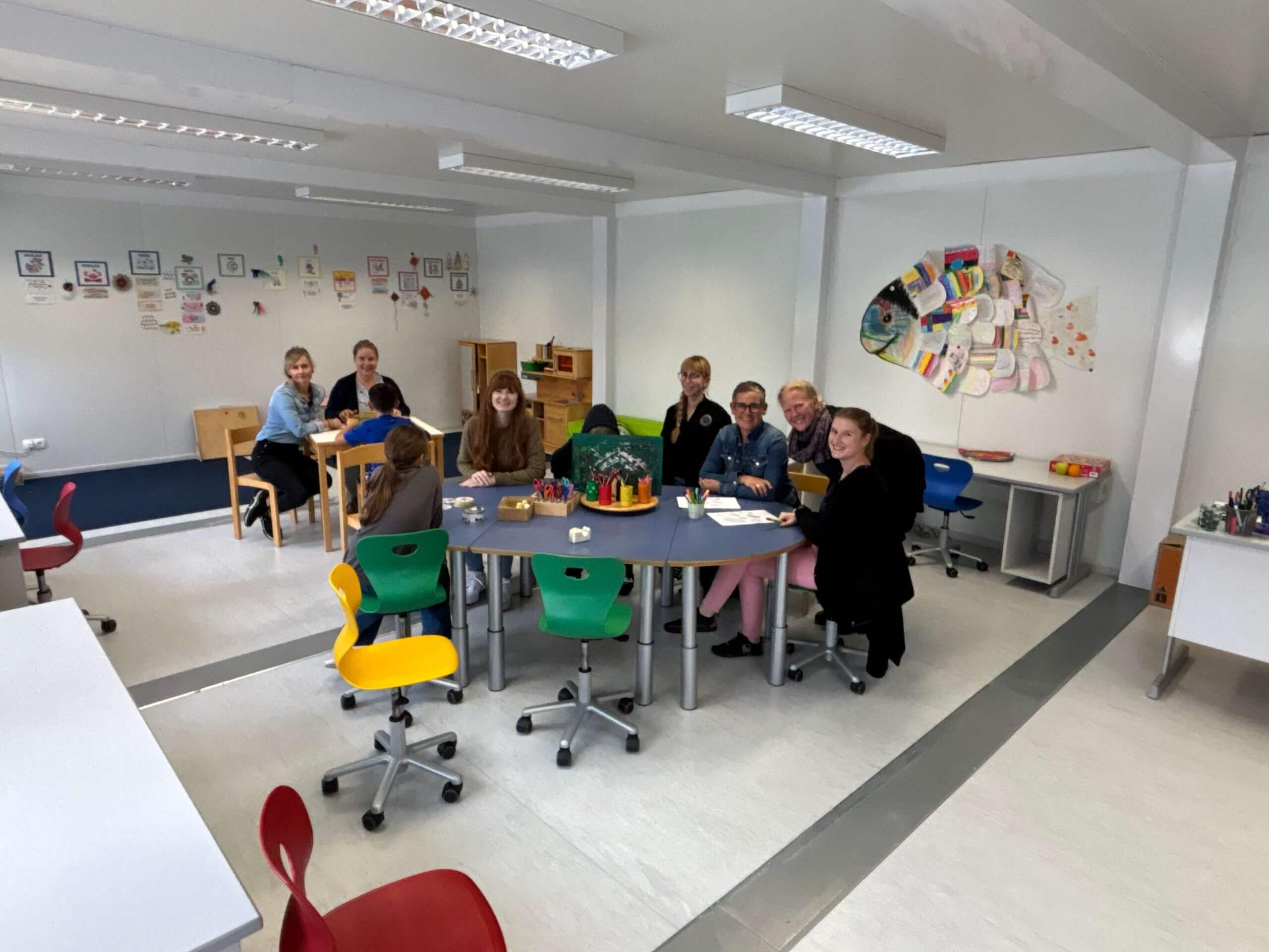 Gruppe von acht Personen, darunter Kinder und Erwachsene, sitzt und arbeitet an mehreren Tischen in einem Klassenraum mit bunten Stühlen und dekorativer Wandgestaltung.