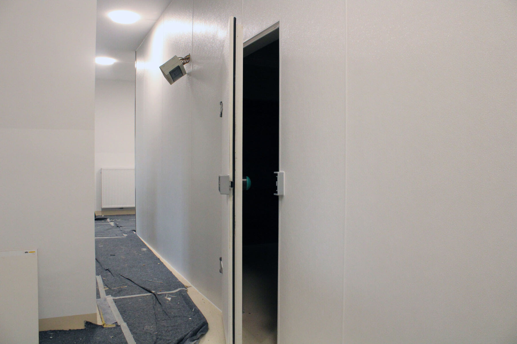 Ein schmaler Flur mit weißen Wänden und einem dunklen Bodenbelag, der teilweise mit Schutzmaterial bedeckt ist. Rechts befindet sich eine offene Tür, die in einen dunkleren Raum führt. An der Wand hängt eine kleine, schräg angebrachte Lampe.