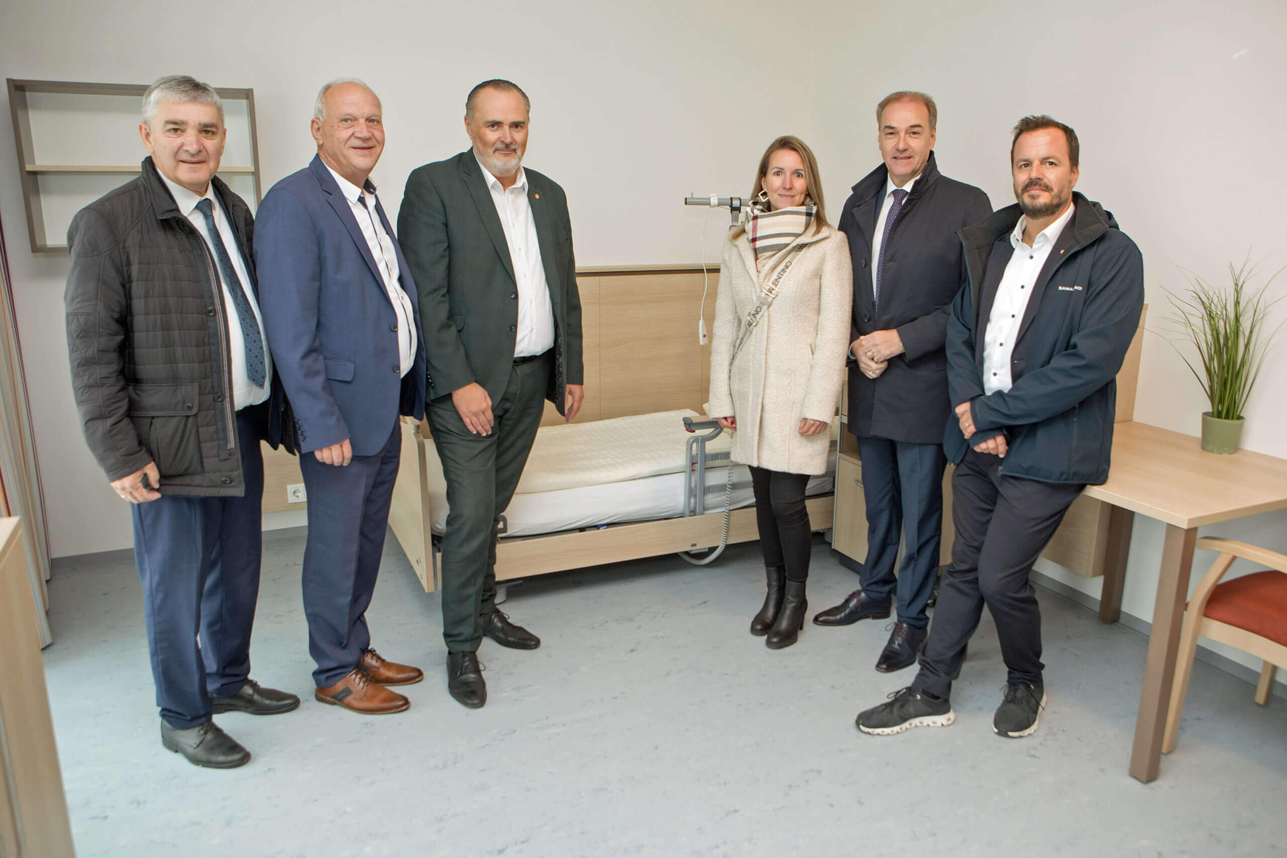 Sechs Personen stehen in einem hellen Raum neben einem Pflegebett mit Holzrahmen. Die Personen tragen formelle Kleidung, die Frau in der Mitte trägt einen beigen Mantel und Schal. Im Hintergrund befindet sich ein Regal und ein Tisch mit einer grünen Zimmerpflanze.