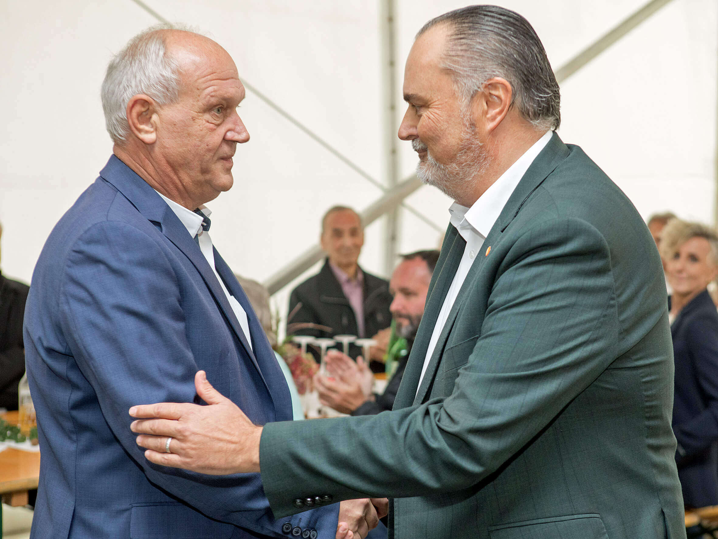 Zwei Männer in Anzügen stehen sich in einem Zelt gegenüber und schütteln sich die Hände. Der Mann rechts trägt einen dunkelgrünen Anzug und hat leicht ergraute Haare. Beide wirken konzentriert. Im Hintergrund sitzen mehrere Personen, die der Szene aufmerksam folgen.