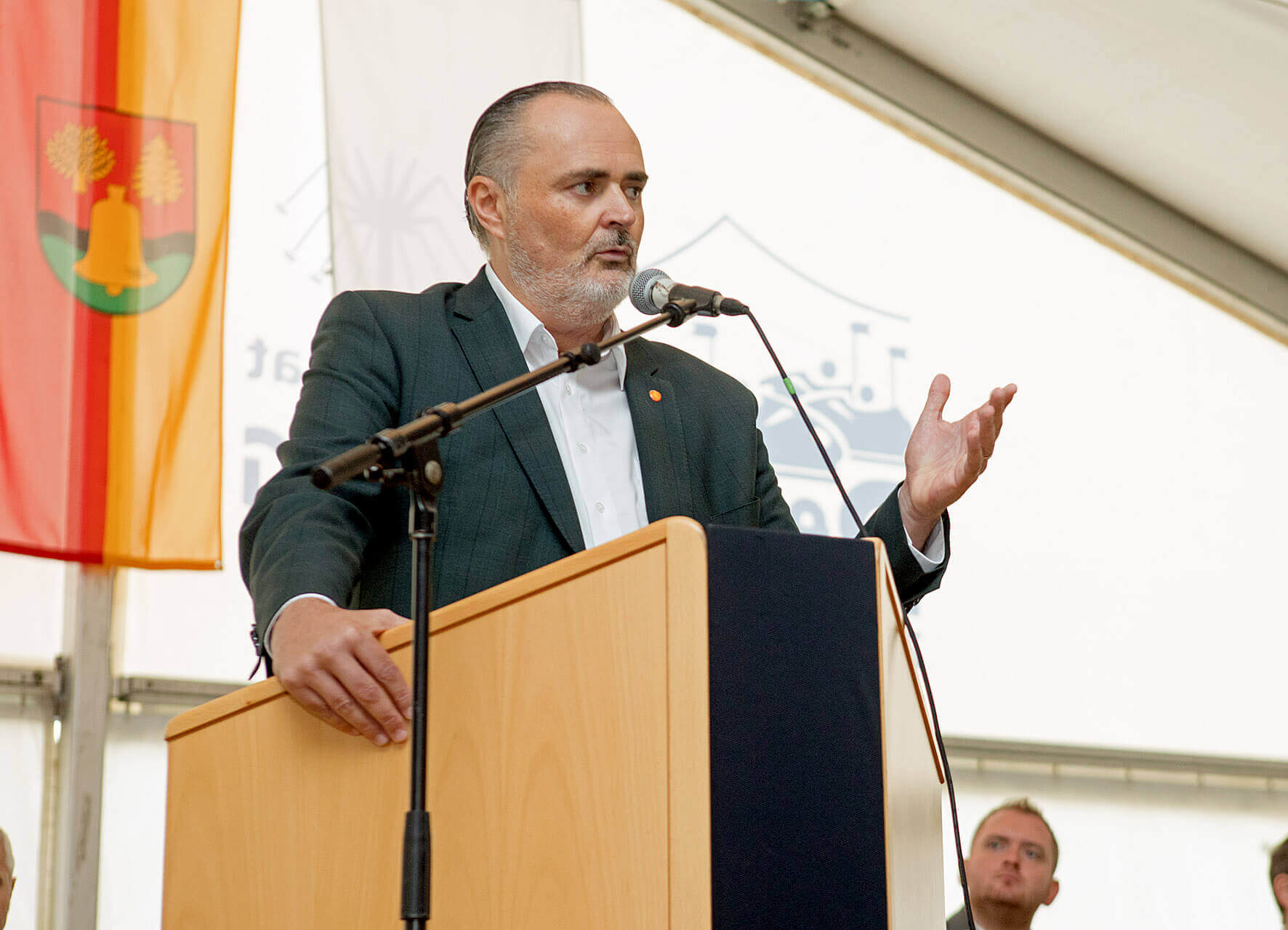 Ein Mann in einem dunkelgrünen Anzug steht an einem Rednerpult und spricht ins Mikrofon. Er gestikuliert mit der rechten Hand und schaut konzentriert ins Publikum. Links neben ihm hängen zwei Fahnen, eine mit einem Wappen, auf dem ein gelbes Glockensymbol abgebildet ist.