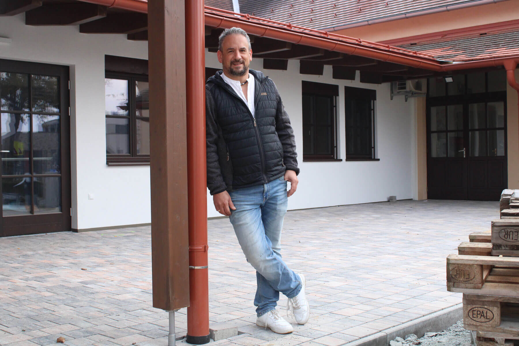 Rene Höfer, ein Mann mit dunkler Jacke und blauer Jeans lehnt an einem braunen Holzpfeiler vor einem Gebäude mit weißen Wänden und großen Fenstern. Der Boden ist mit grauen Pflastersteinen gepflastert, und rechts im Bild sind gestapelte Holzpaletten zu sehen.