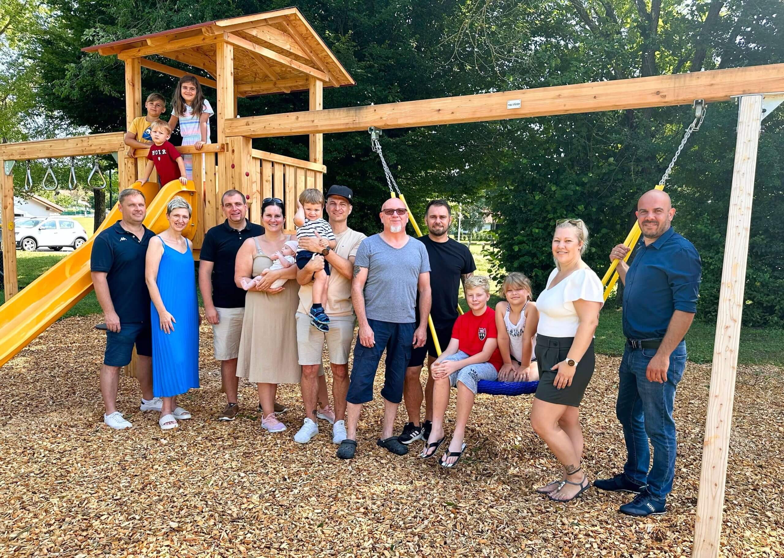 Ein Gruppenfoto von 15 Personen, darunter Männer, Frauen und Kinder, die vor einem Holzspielgerüst mit gelber Rutsche stehen. Im Vordergrund liegt der Boden mit Holzschnitzeln bedeckt, und im Hintergrund sind grüne Bäume zu sehen. Die Gruppe lächelt in die Kamera, und die Stimmung wirkt familiär und entspannt.