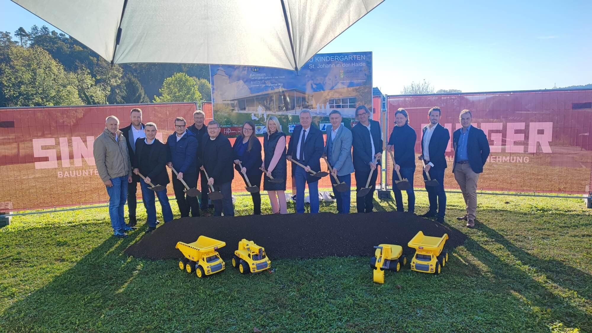 Eine Gruppe von etwa zwölf Personen steht vor einem aufgehäuften Erdhügel auf einer grünen Wiese. Sie halten Schaufeln in den Händen, als ob sie gerade einen symbolischen Spatenstich durchführen. Vor dem Hügel sind vier gelbe Spielzeugbaumaschinen zu sehen (darunter Bagger und Kipper). Im Hintergrund ist ein Bauzaun mit dem Schriftzug "Singer Bauunternehmung" sowie ein großes Schild mit der Aufschrift „Zu- und Umbau Kindergarten St. Johann in der Haide“ zu erkennen. Der Tag ist sonnig.