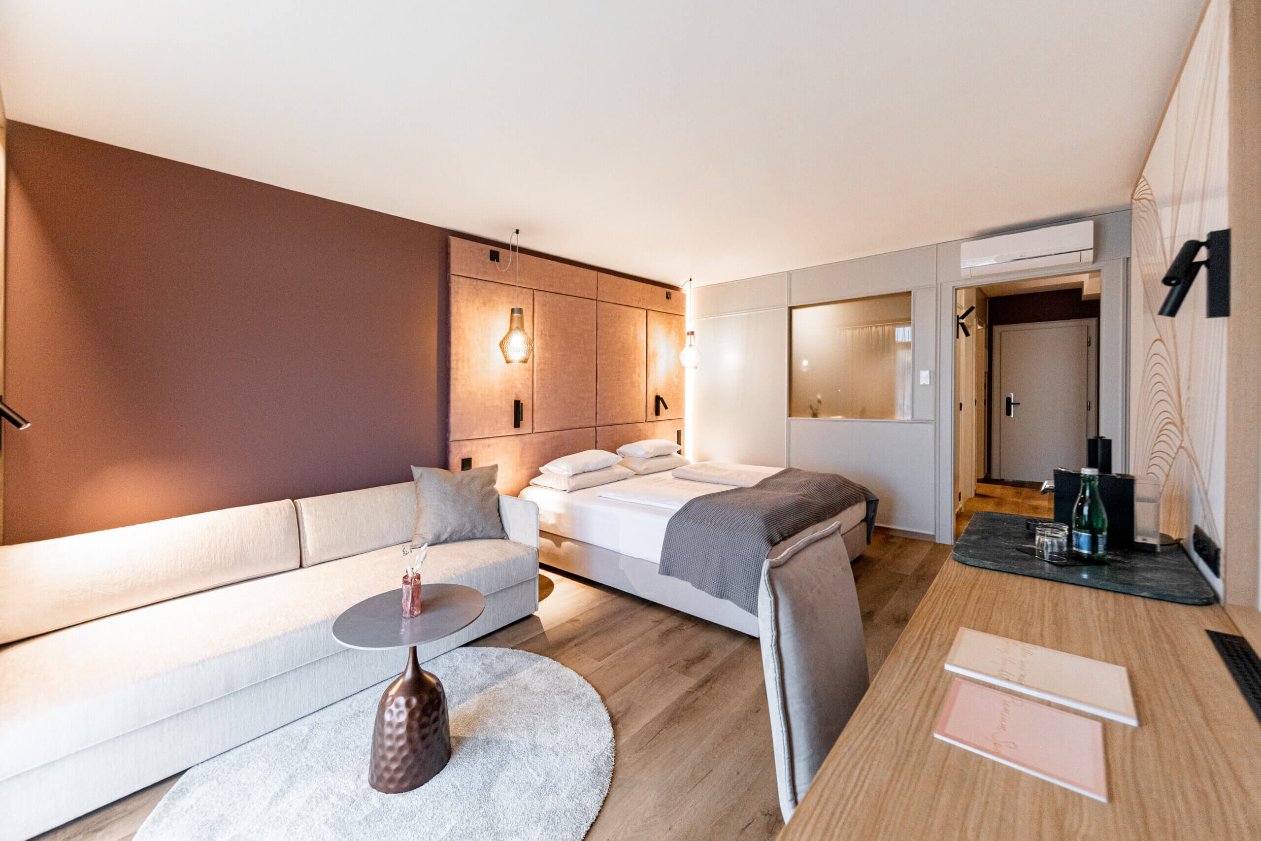 Ein modernes Wellnesszimmer im AVITA Resort. Das Zimmer ist geschmackvoll eingerichtet mit einem großen Bett, einem Sofa, einem runden Couchtisch und einer stilvollen Holzanrichte. Die warmen Farben und die Kombination aus Holz, Stoffen und Licht schaffen eine gemütliche und einladende Atmosphäre. Eine Fensterscheibe trennt das Schlafzimmer optisch von einem angrenzenden Badezimmerbereich.