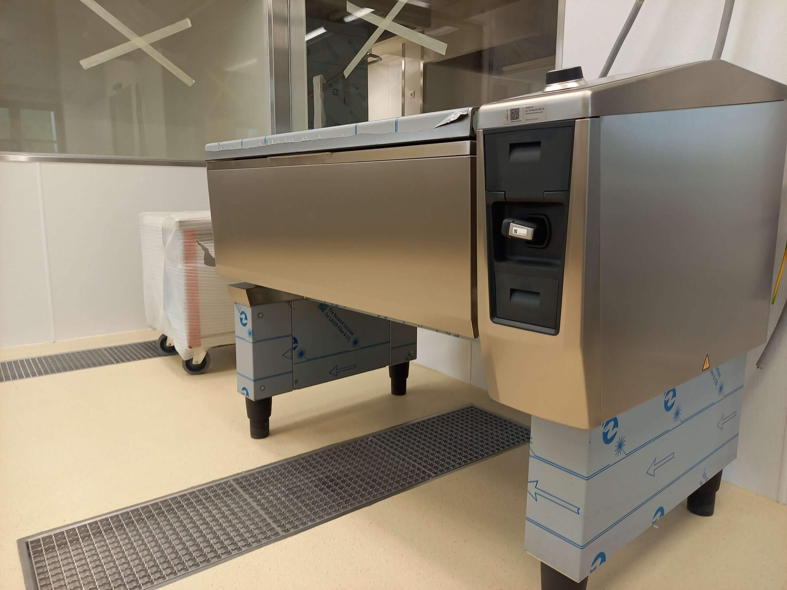 Detailaufnahme einer Edelstahlkücheneinrichtung in einem hellen Raum mit beigem Boden und Gitterabläufen. Die Geräte haben eine glänzende Metalloberfläche, und links steht ein mit Folie umwickelter Rollcontainer vor einer Glasabtrennung.