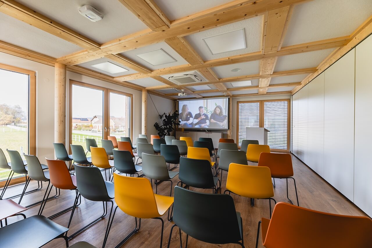 Moderner Seminarraum mit bunten Stühlen und Leinwand, helles Tageslicht durch große Fenster. Ideal für Meetings und Präsentationen.