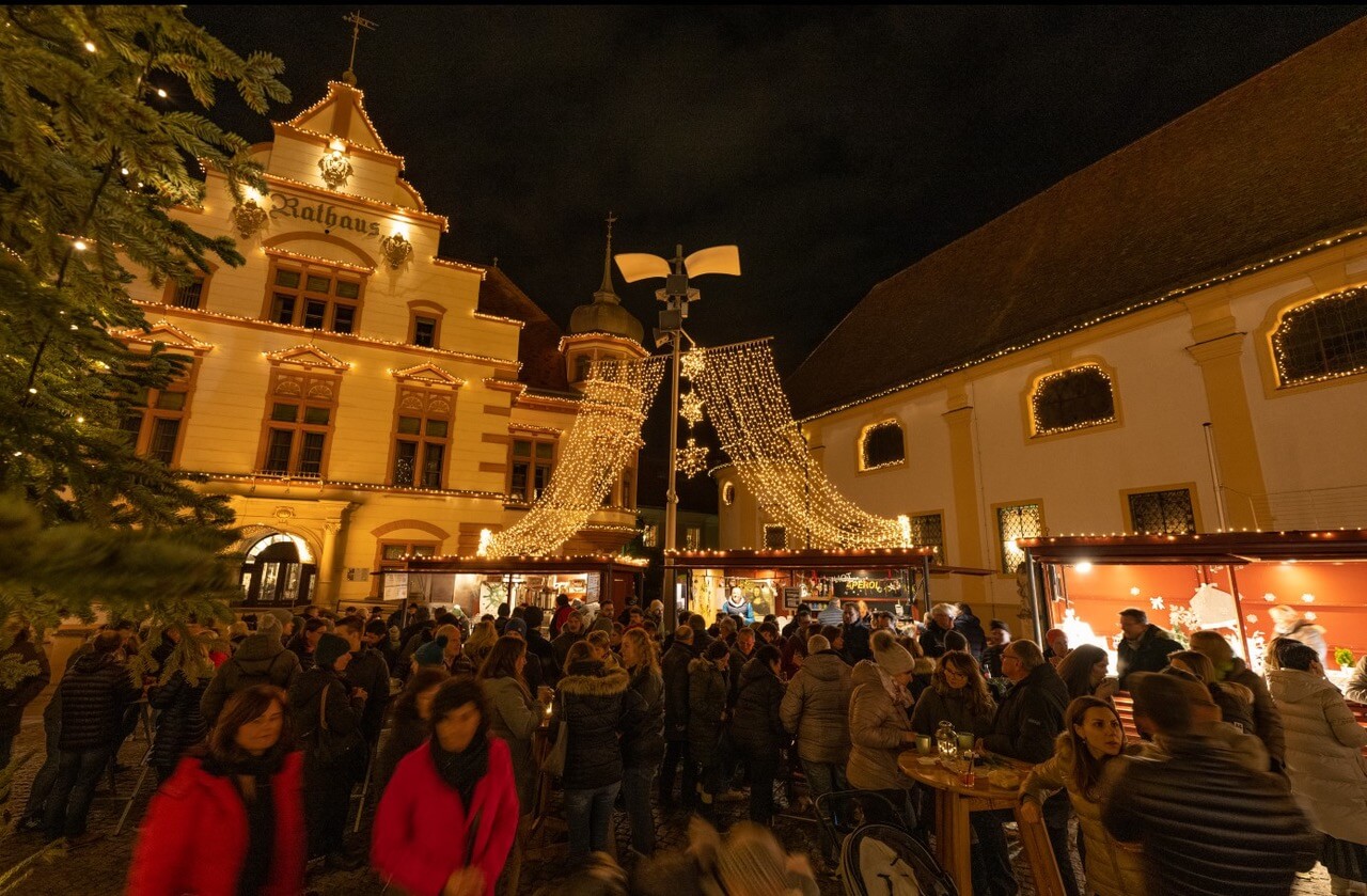 Weihnachtsmarkt bei Nacht, beleuchtetes Rathaus mit Menschenmenge im festlichen Ambiente.