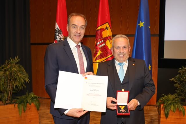 Zwei Männer bei einer Preisverleihung mit Medaille und Urkunde vor europäischen und nationalen Flaggen.