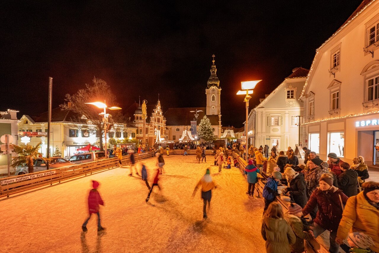 Nachtszene auf einem Weihnachtsmarkt mit Eislaufbahn und beleuchteter Kirche im Hintergrund, Menschen genießen die Winterstimmung.
