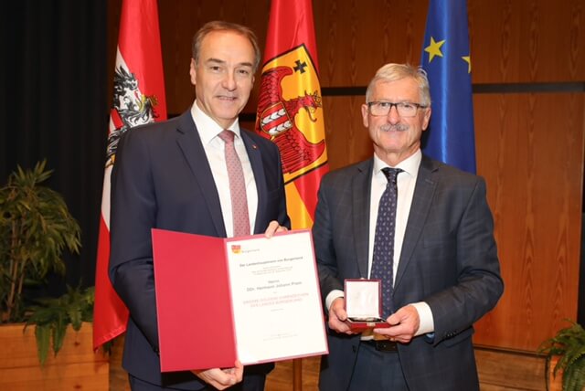 Zwei Männer bei einer Preisverleihung, eine Urkunde und eine Medaille vor Flaggen im Hintergrund.