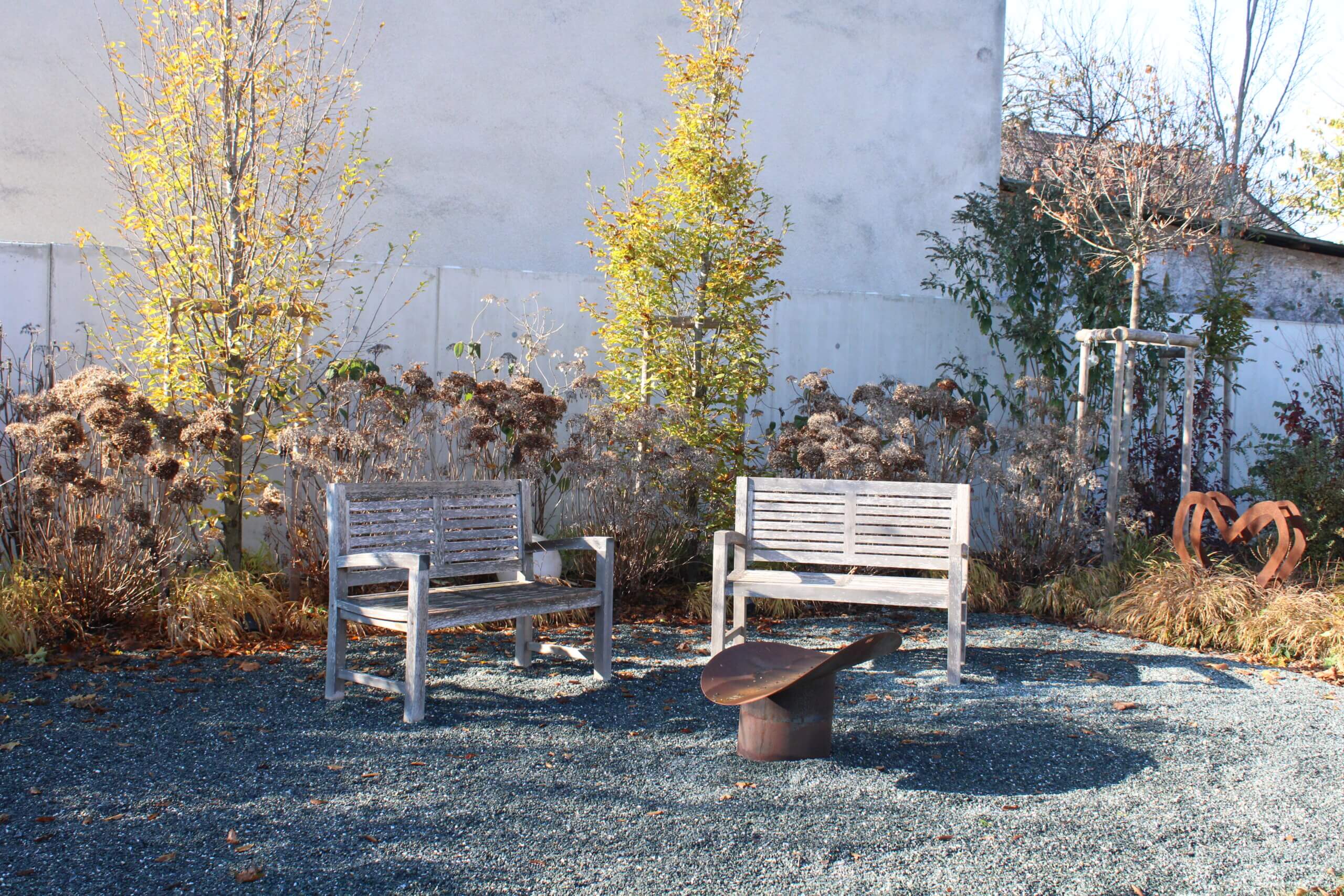 Zwei Holzbänke im herbstlichen Garten mit Feuerstelle und dekorativer Skulptur auf Kiesboden.
