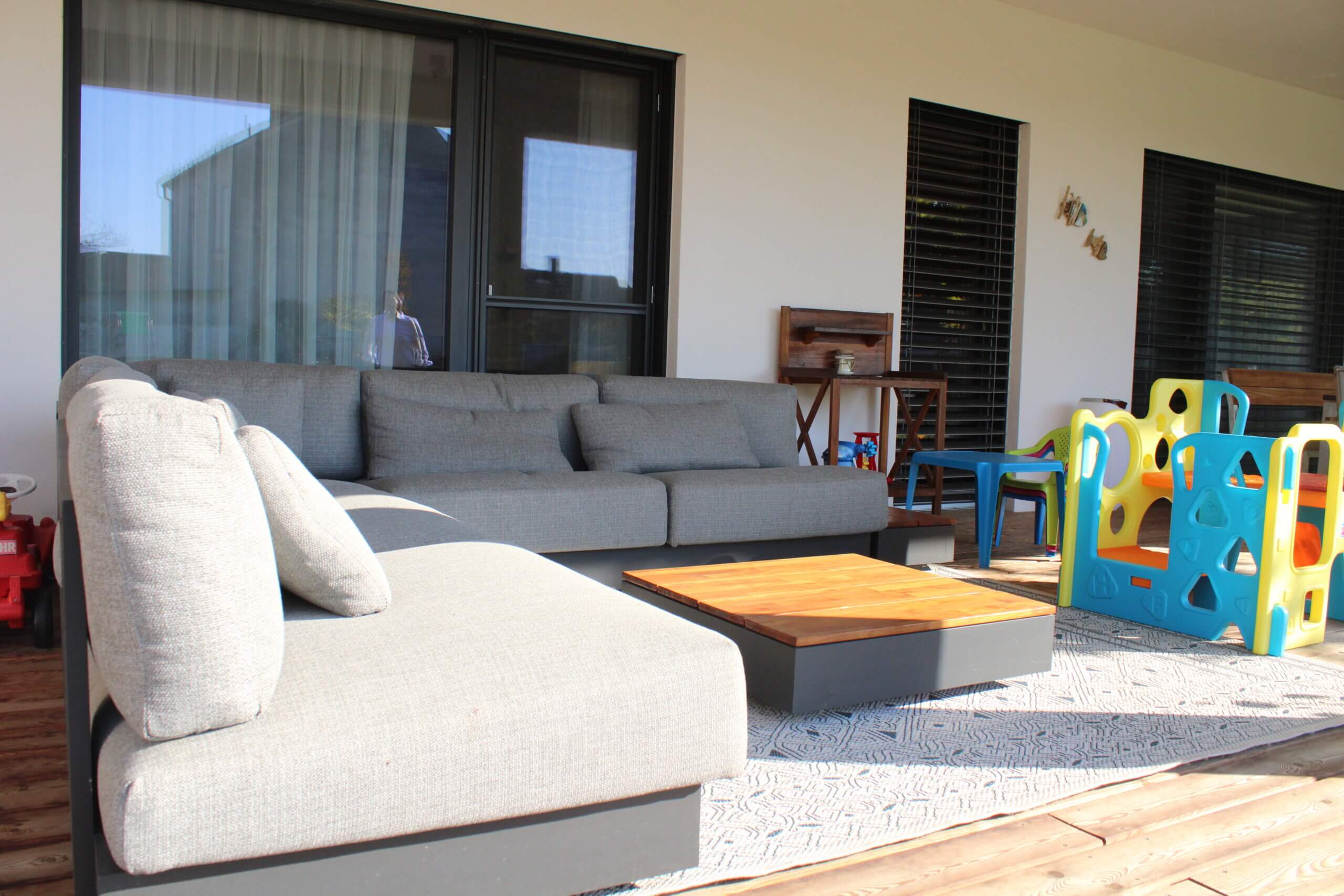 Moderne Terrasse mit grauer Couch und Kinderspielzeug, ideal für Familien. Gemütlicher Außenbereich bei Sonnenschein.