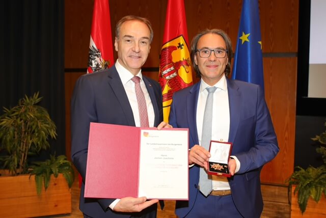 Zwei Männer bei einer Preisverleihung, einer hält eine Urkunde, der andere eine Medaille, vor Flaggenhintergrund.