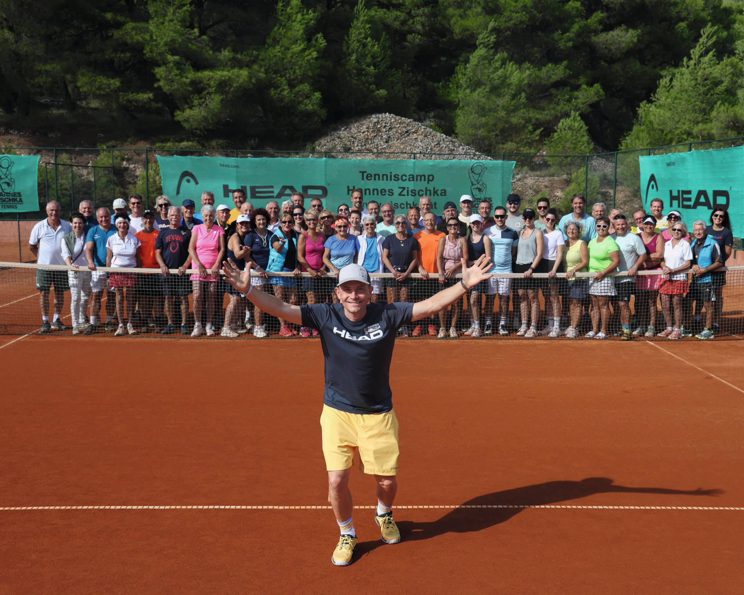 Gruppe von Tennisspielern posiert auf Sandplatz während eines Tenniscamps bei sonnigem Wetter.