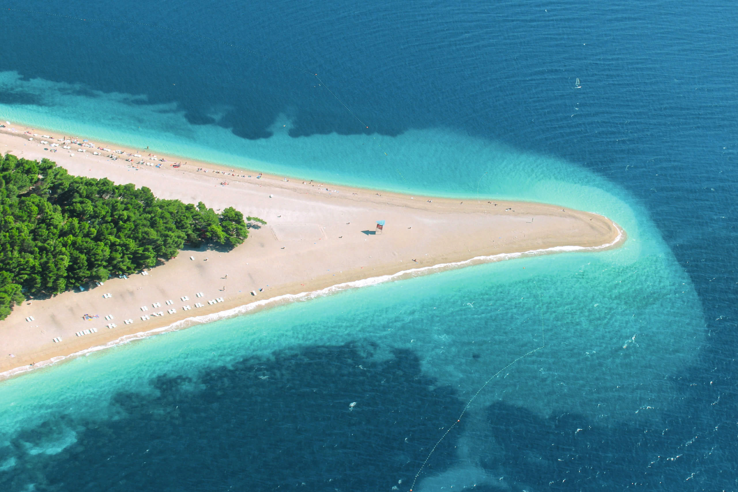Luftaufnahme des Zlatni Rat Strandes in Kroatien mit klarem, türkisfarbenem Wasser und Pinienwald entlang der Küste.