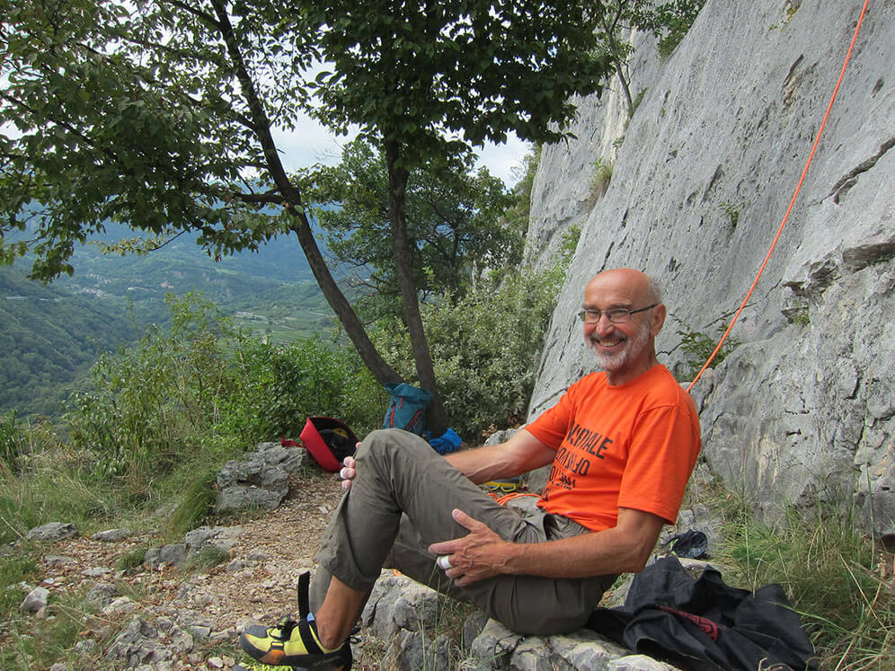 Mann entspannt beim Klettern in den Bergen, lachend auf Felsen sitzend, mit Seil und Ausrüstung in malerischer Naturumgebung.