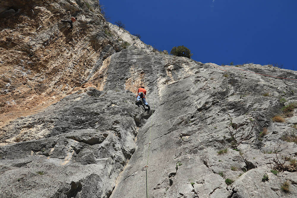 Kletterer in orangeem Shirt erklimmt steile Felswand unter blauem Himmel, symbolisiert Abenteuer und Entschlossenheit.