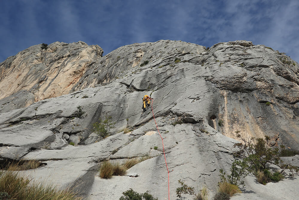 Kletterer in gelber Ausrüstung am Felsen unter blauem Himmel, Aufstieg in natürlicher Berglandschaft.
